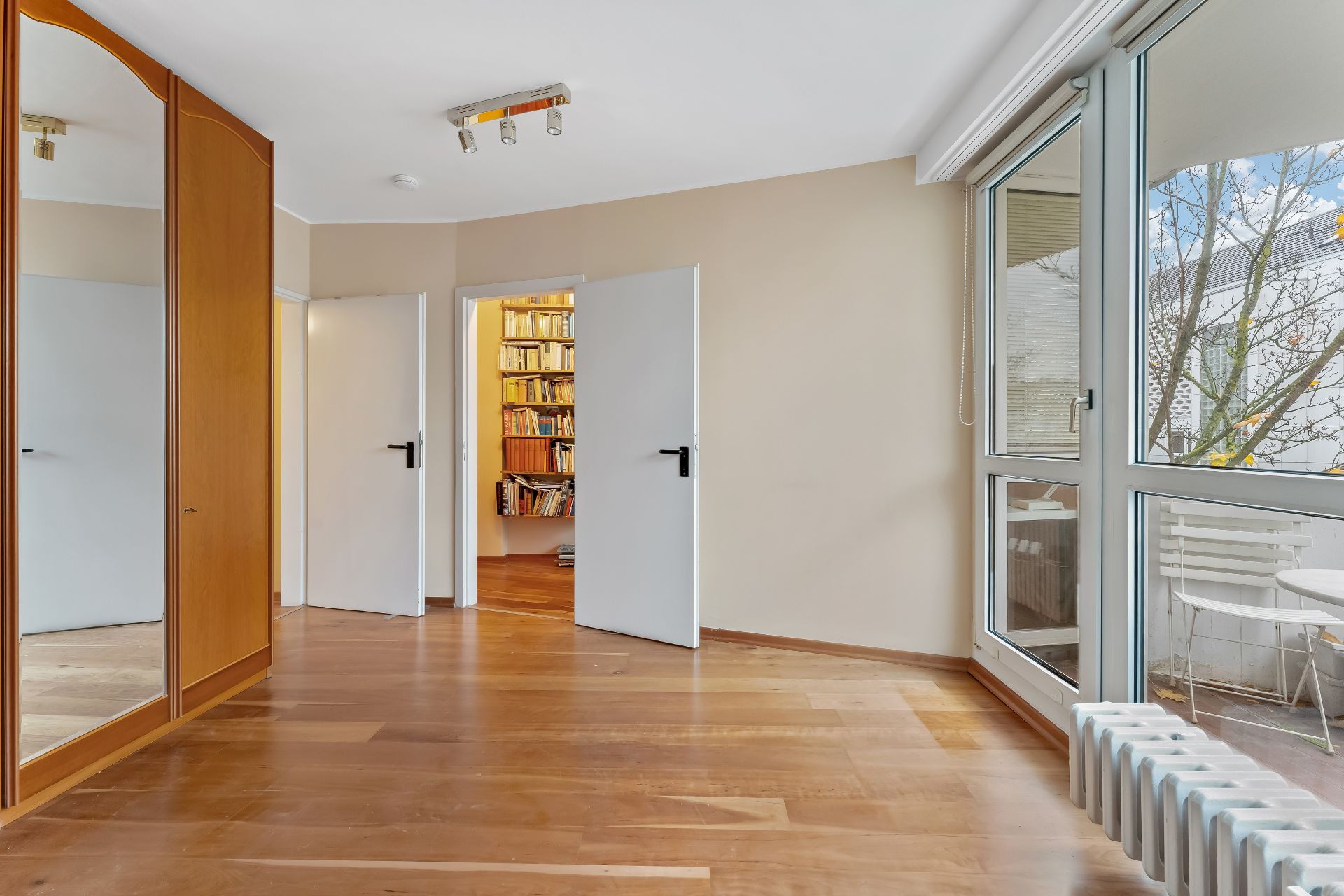 Helles Zimmer mit Holzfußboden, Einbauschrank, großen Fenstern und Blick auf Bücherregal im Nebenraum.