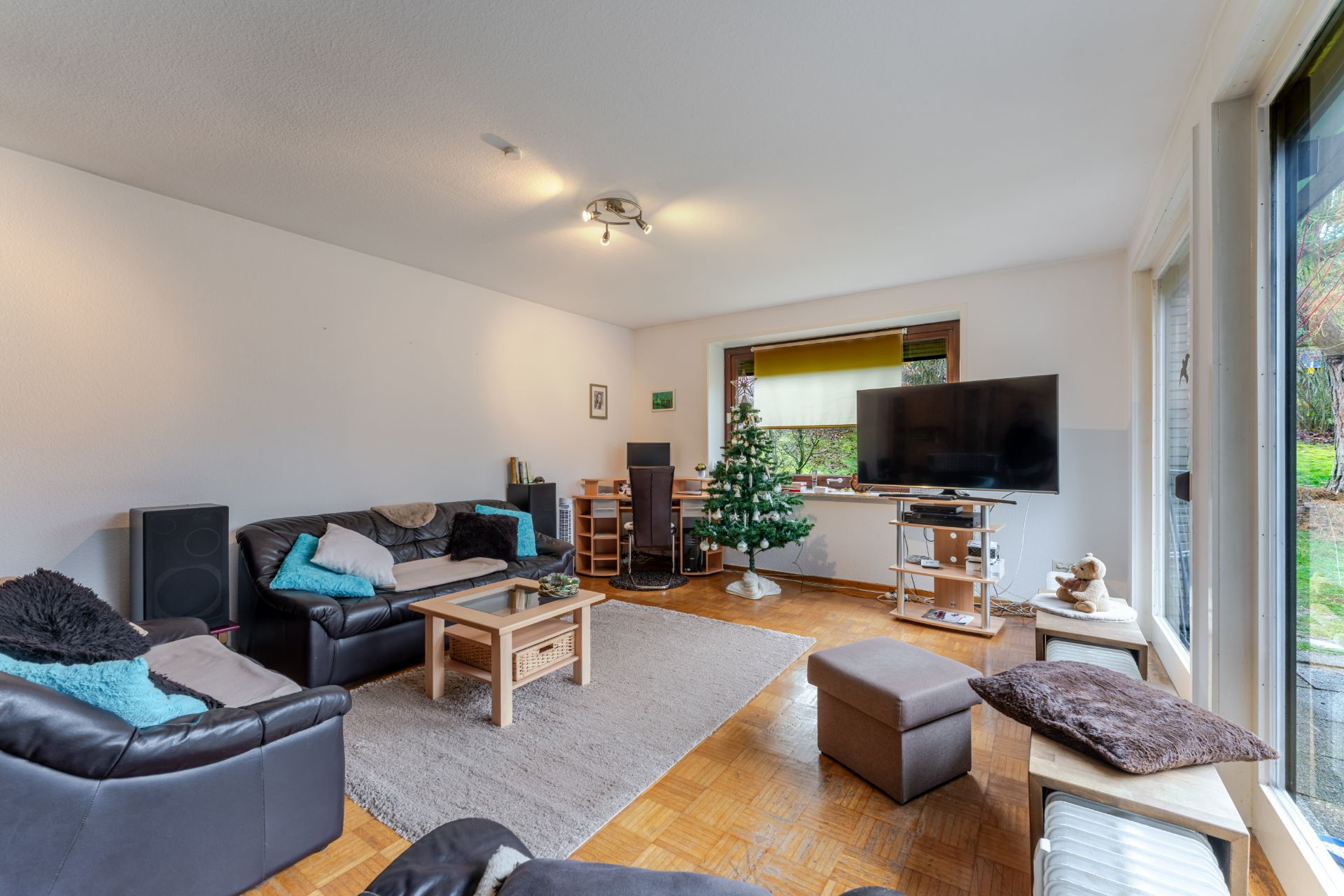 Wohnzimmer mit Sofas, Fernseher, Couchtisch und kleinem Weihnachtsbaum, große Fenster zum Garten.