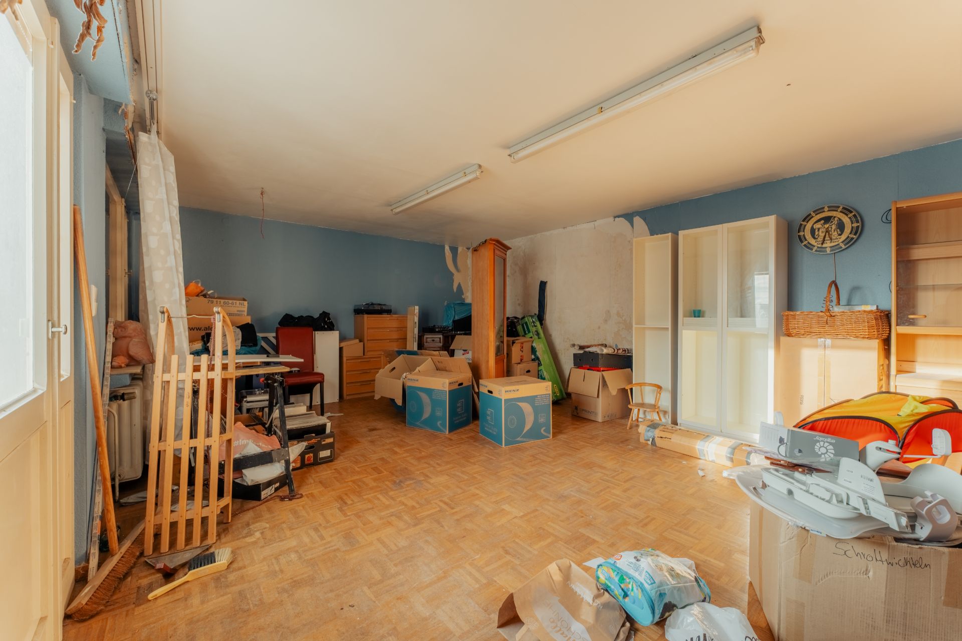 Ein großes, unaufgeräumtes Zimmer mit Holzboden, Kartons und verschiedenen Möbelstücken.