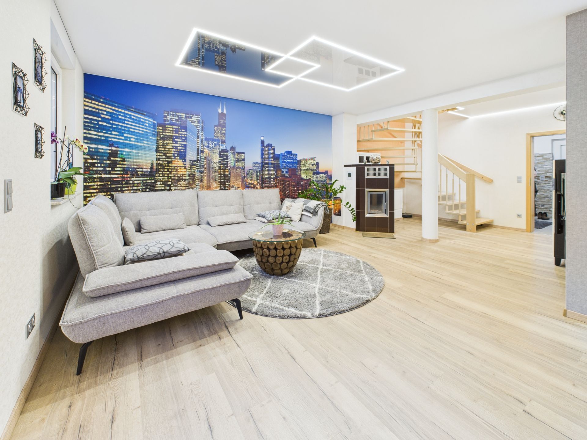 Modernes Wohnzimmer mit Ecksofa, rundem Teppich und Stadtbild-Wandbild, daneben eine offene Treppe.