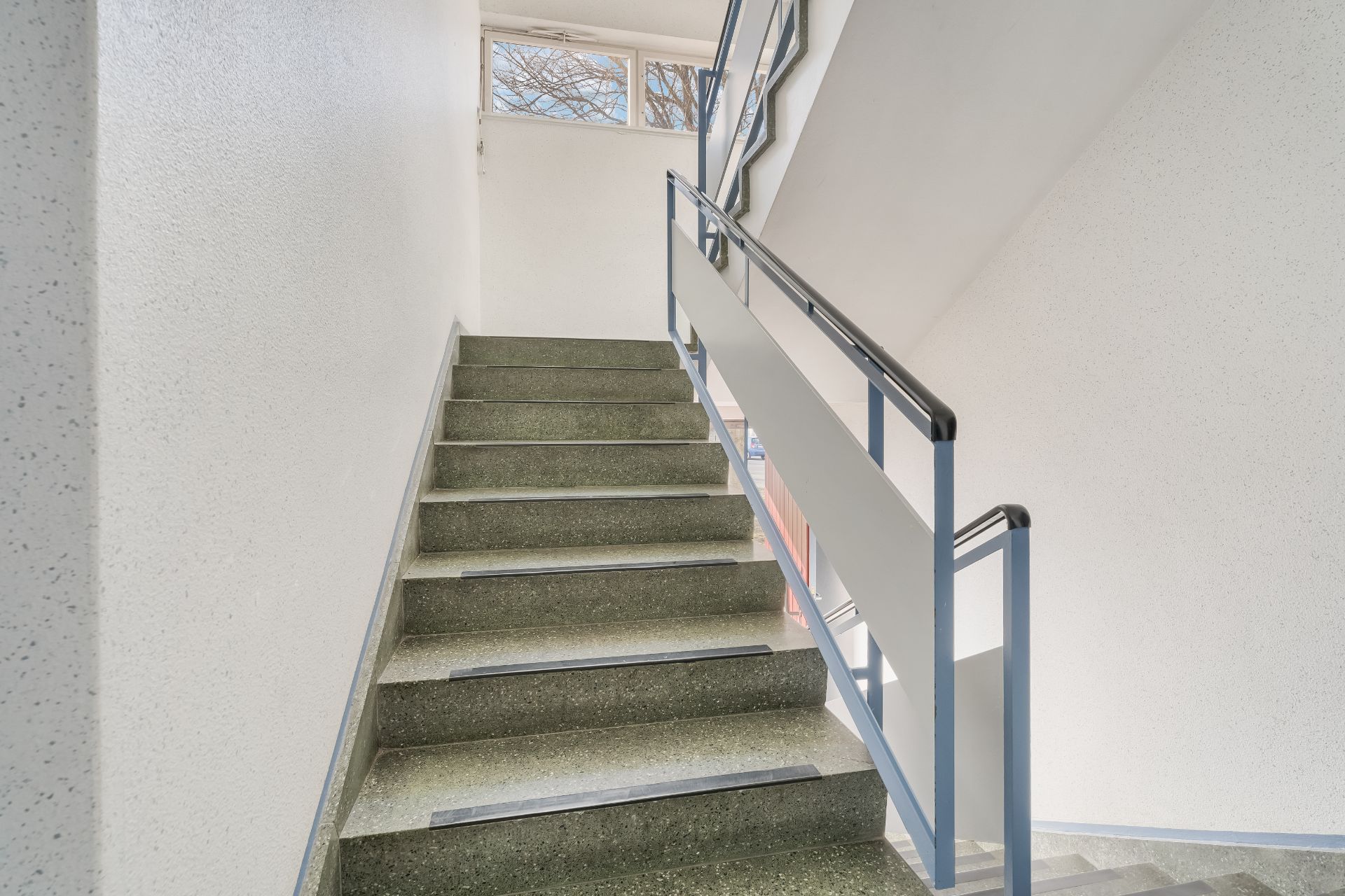 Helles Treppenhaus mit grünlichen Stufen und Metallgeländer, Fenster oben.