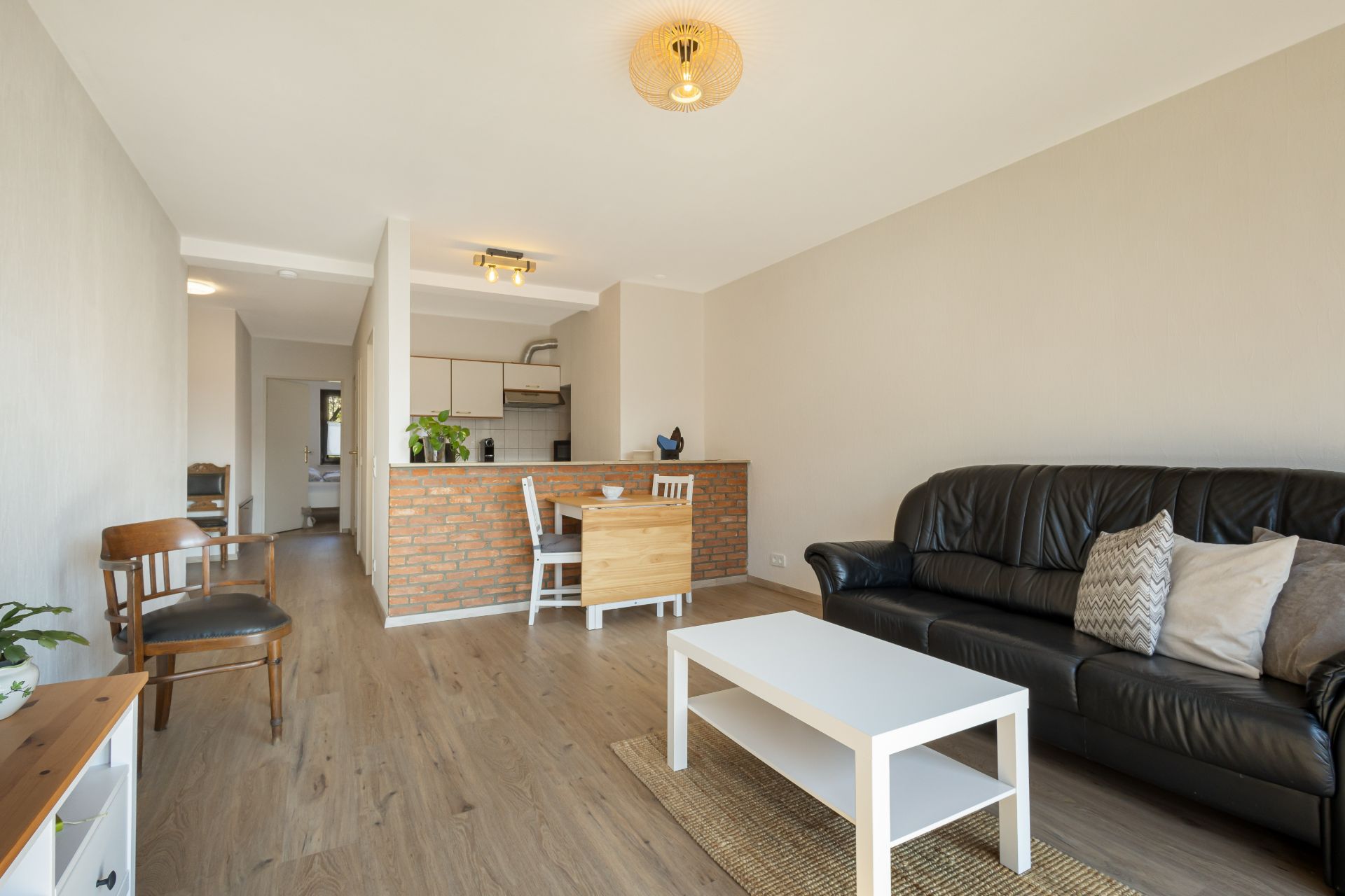 Helles Wohnzimmer mit schwarzem Sofa, weißem Tisch und offener Küche mit Essbereich.