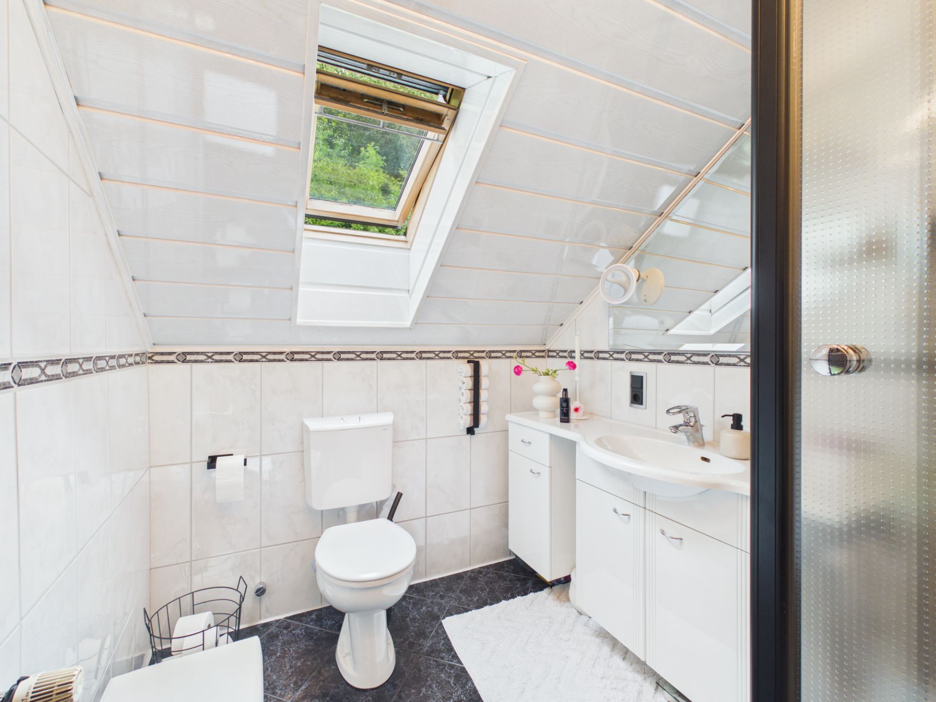 Helles Badezimmer mit Dachfenster, Toilette, Waschbecken und Dusche unter einer schrägen Decke.