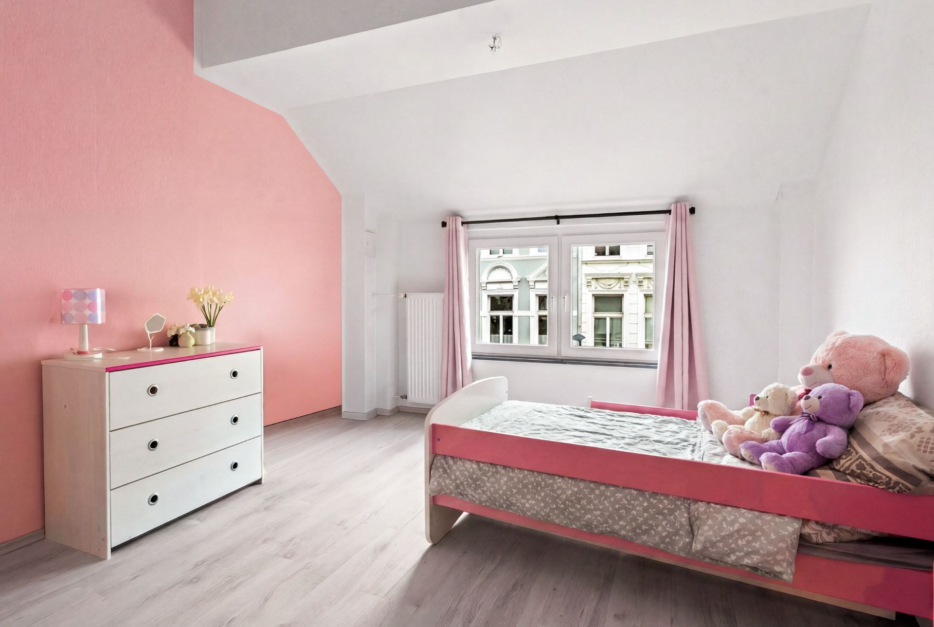 Helles Kinderzimmer mit rosa Wand, Einzelbett, Kuscheltieren und Kommode vor einem Fenster.