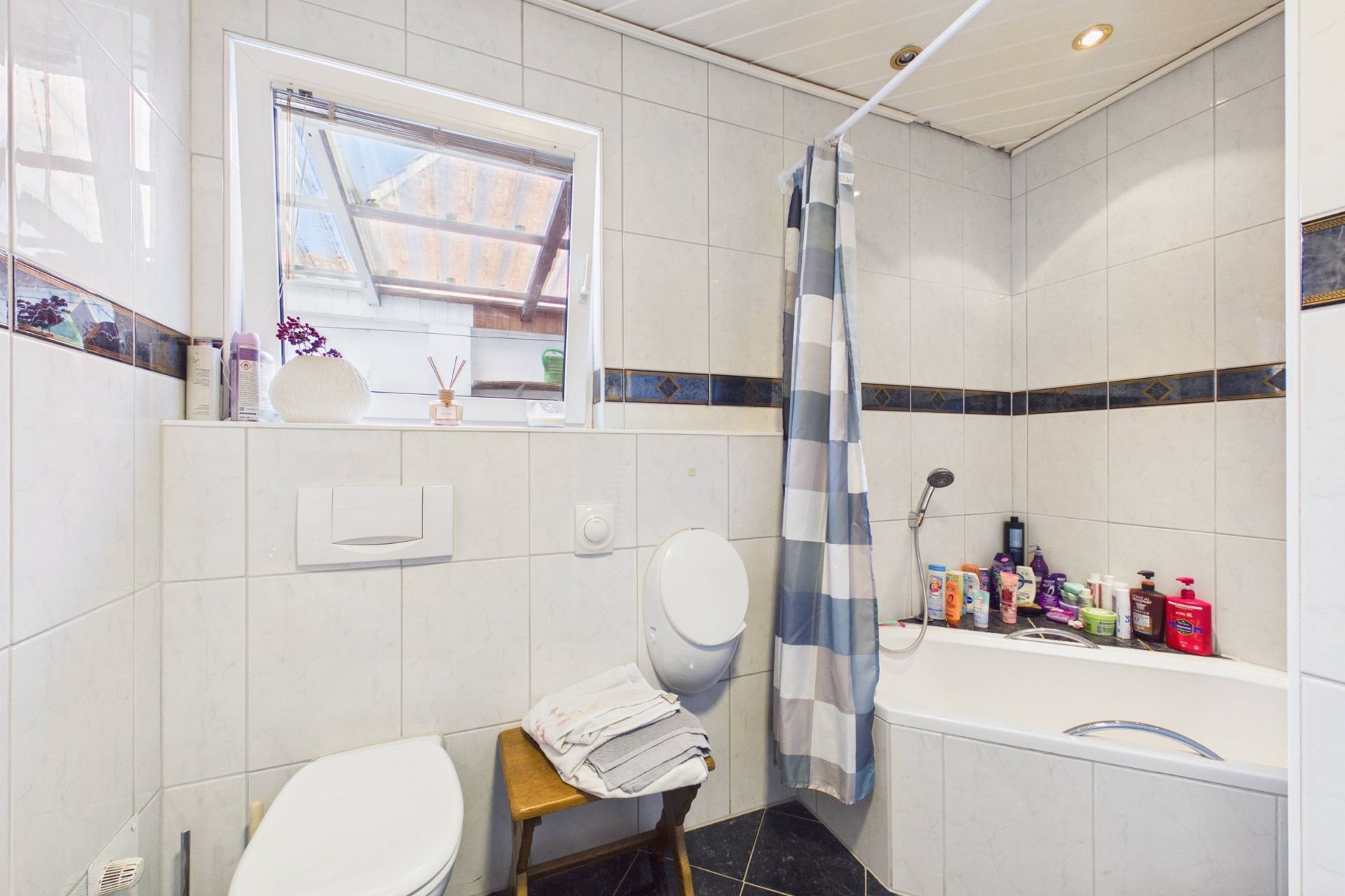 Badezimmer mit Badewanne, Duschvorhang, Toilette und Fenster, einige Pflegeprodukte sind sichtbar.