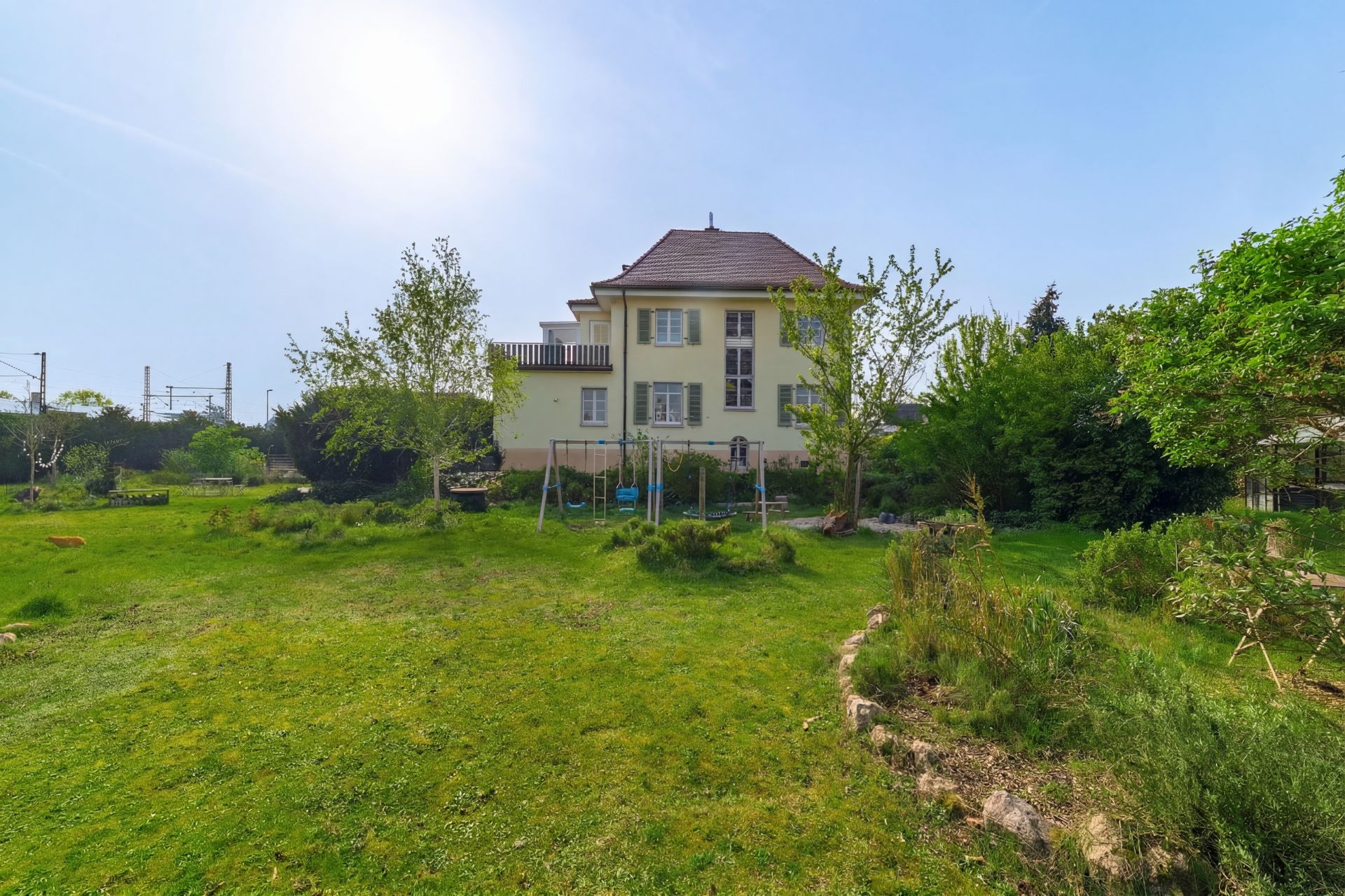 Gelbes Wohnhaus mit großem Garten und Bäumen im Vordergrund an einem sonnigen Tag.