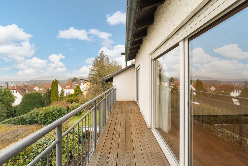 Langer Balkon mit Holzboden und Glasgeländer, Blick auf Nachbarhäuser und Garten.