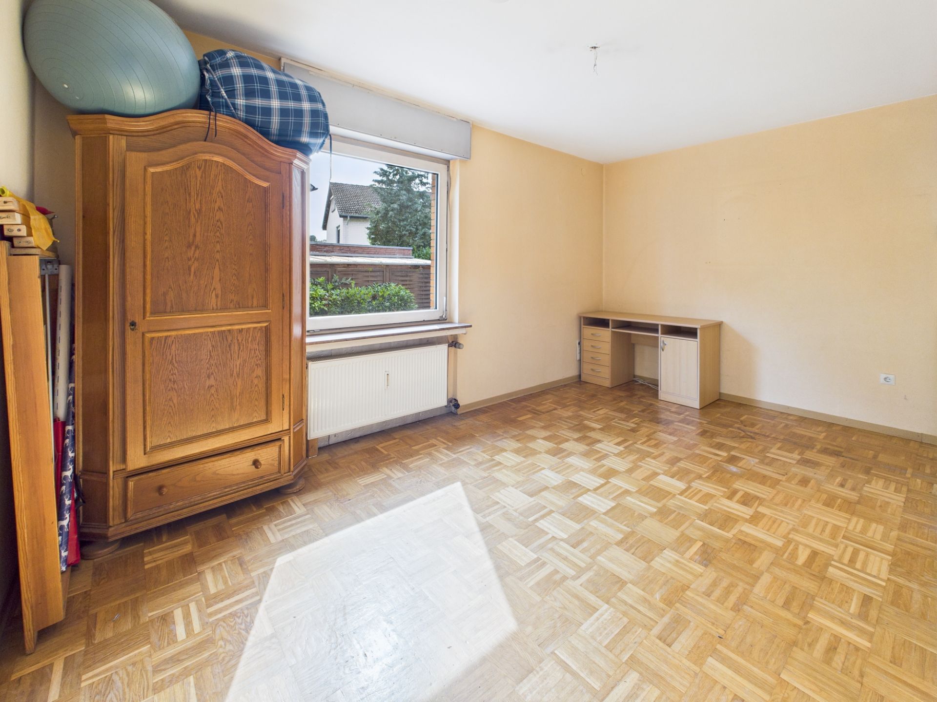 Helles Zimmer mit Parkettboden, großem Fenster, Holzschrank und Schreibtisch.