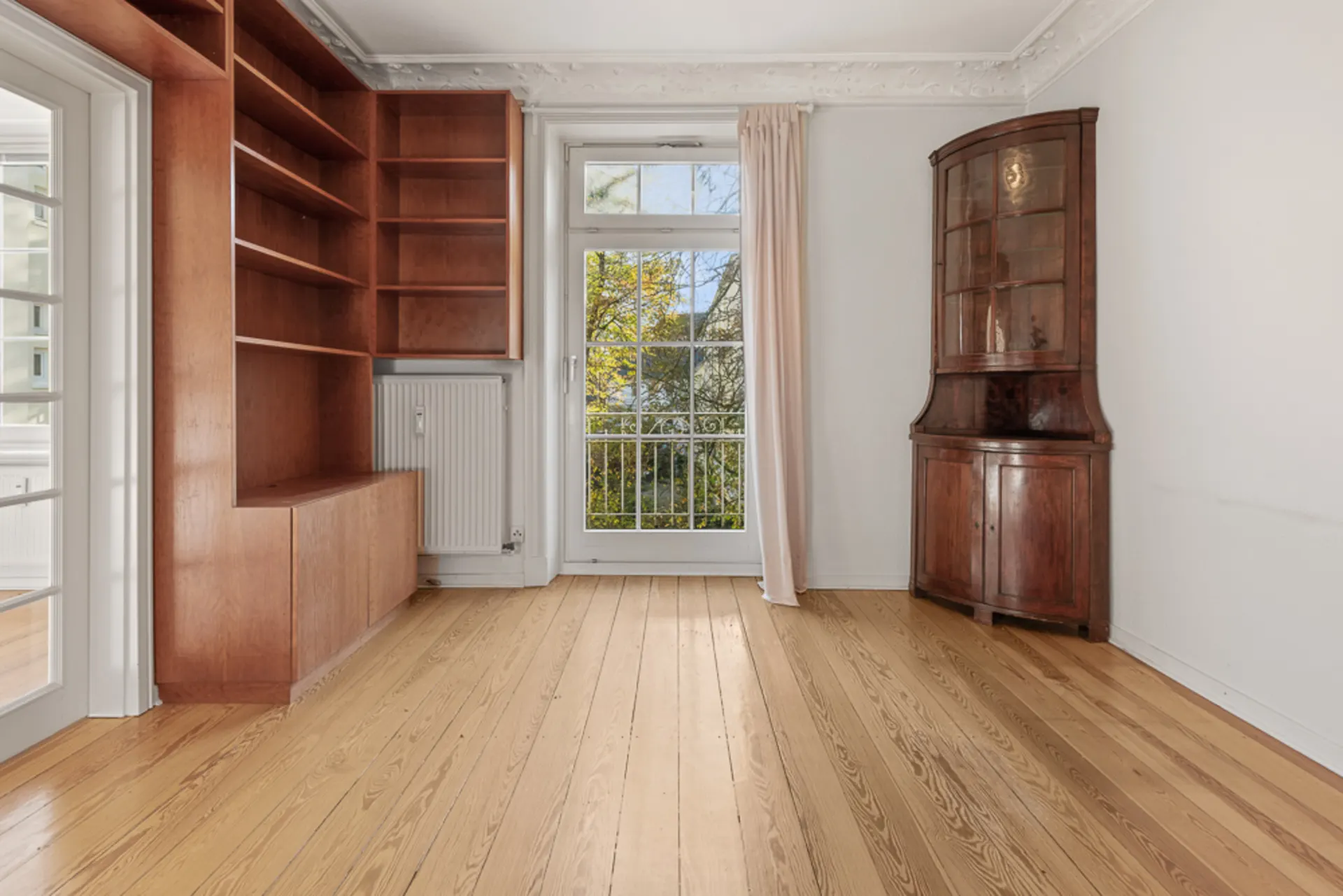 Zimmer mit Holzboden, Regalwand, Glastür, Fenster und Eckschrank.