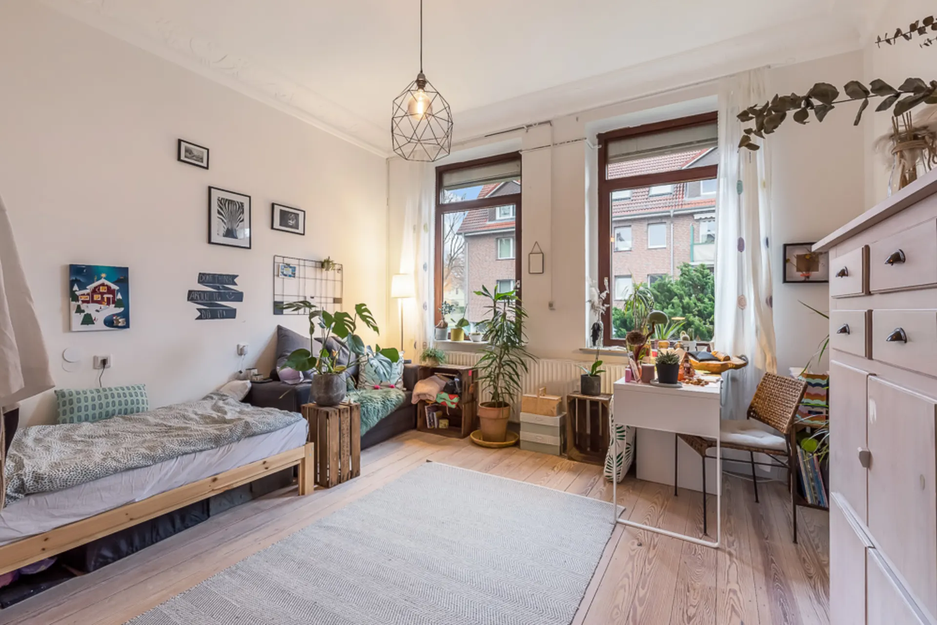 Helles Zimmer mit Bett, Sofa, Schreibtisch, Pflanzen und großen Fenstern.