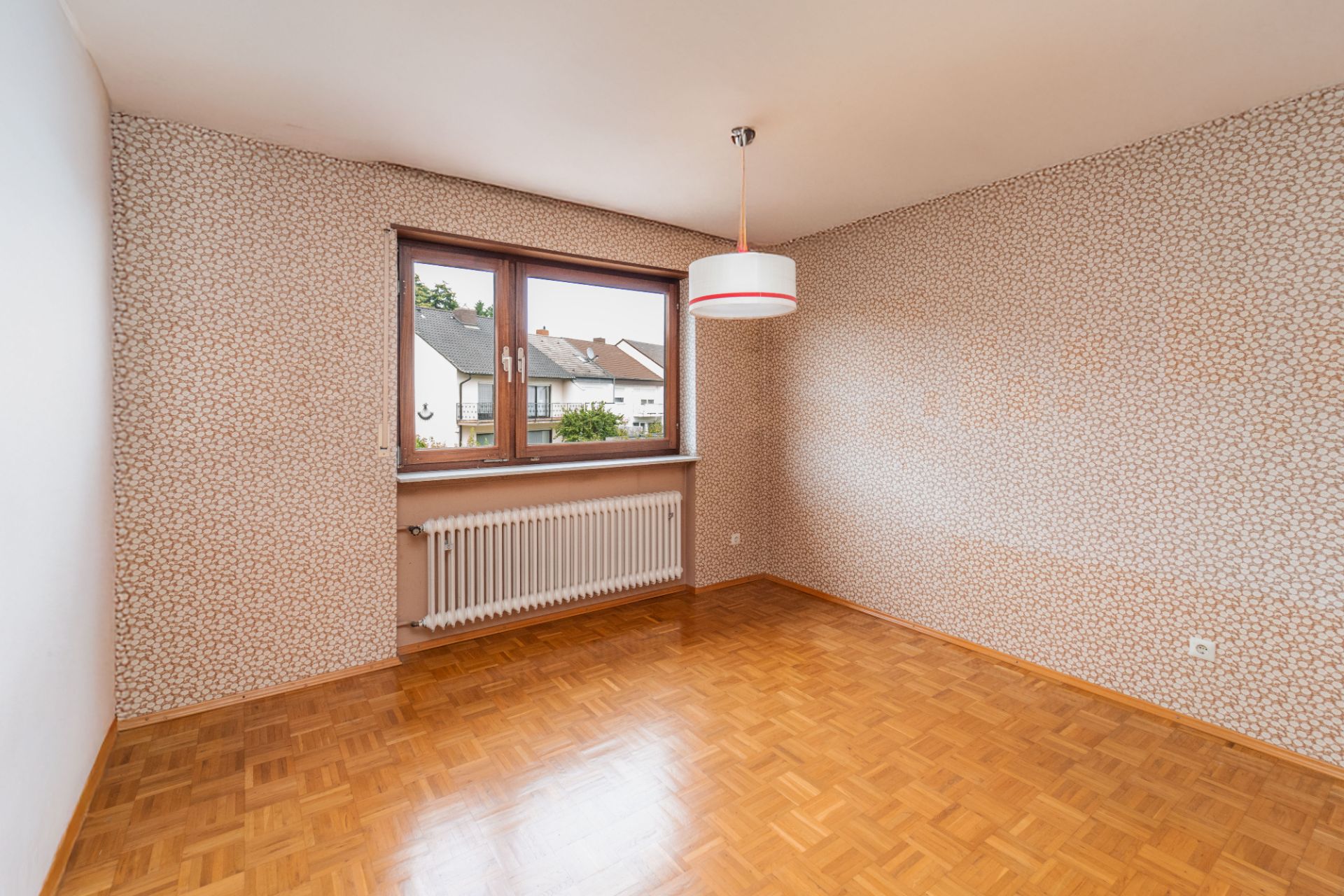 Leeres Zimmer mit Parkettboden, gemusterter Tapete, Fenster, Heizkörper und Deckenlampe.