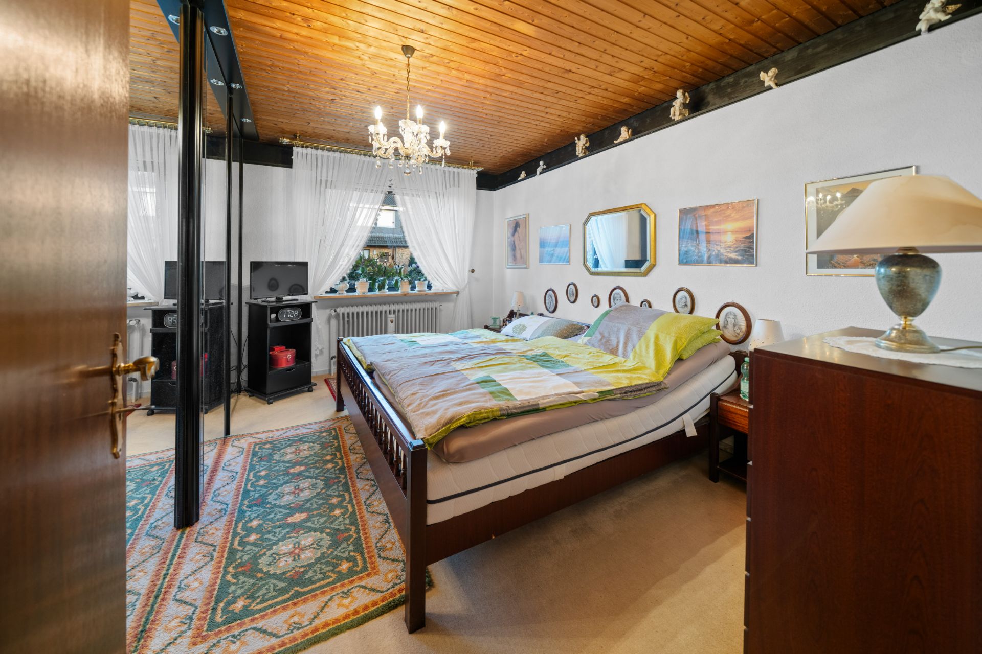 Schlafzimmer mit Doppelbett, Holzdecke, Teppich, Spiegel und Fenster mit weißen Vorhängen.