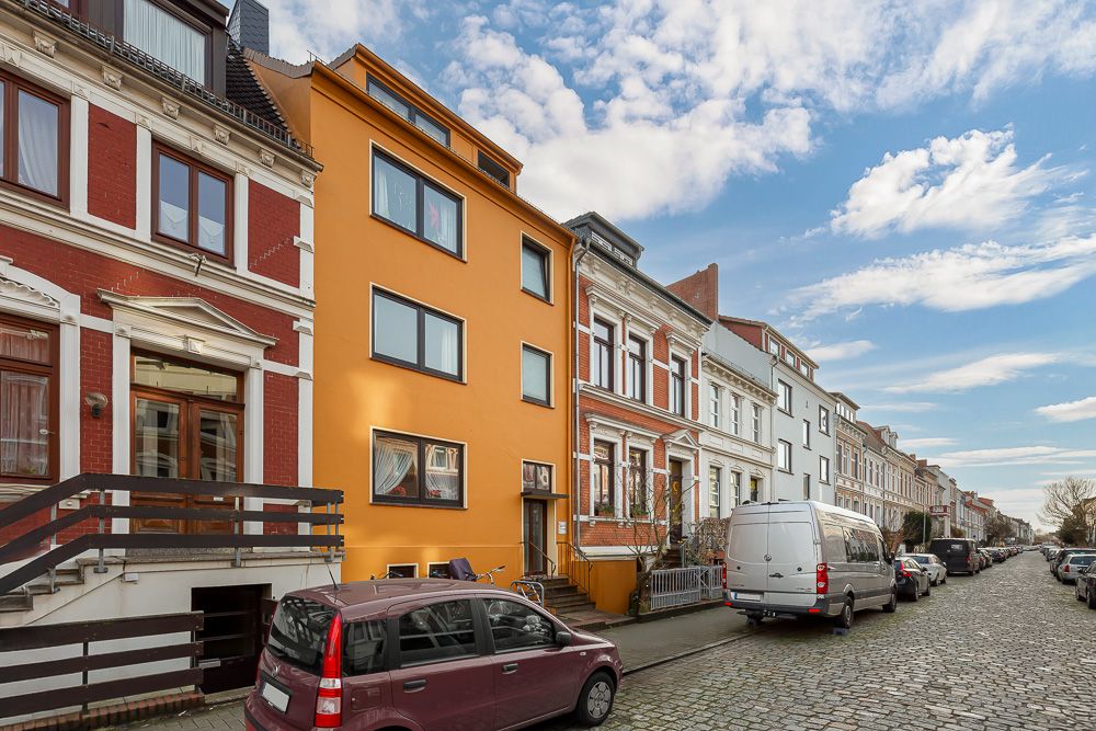 Mehrfamilienhäuser mit bunten Fassaden und parkenden Autos an einer Kopfsteinpflasterstraße.