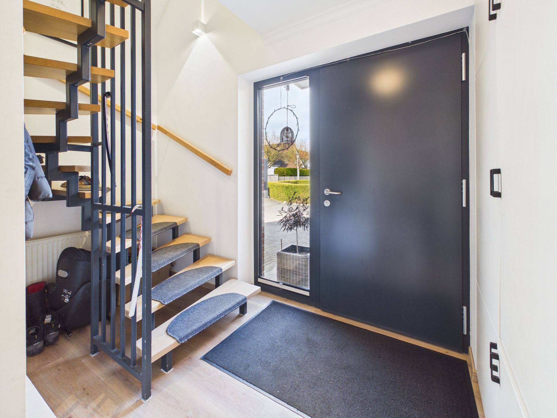 Heller Eingangsbereich mit dunkelgrauer Tür, Treppe aus Holz und Metall, sowie einer Fußmatte.