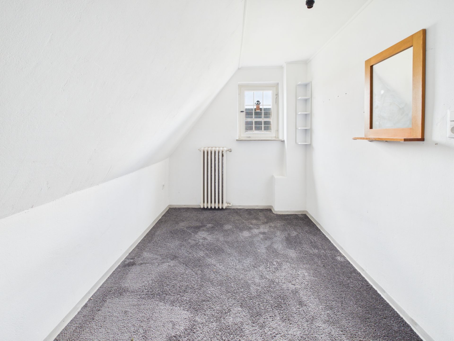 Kleines Zimmer mit grauem Teppich, kleinem Fenster, Heizkörper und Wandspiegel.