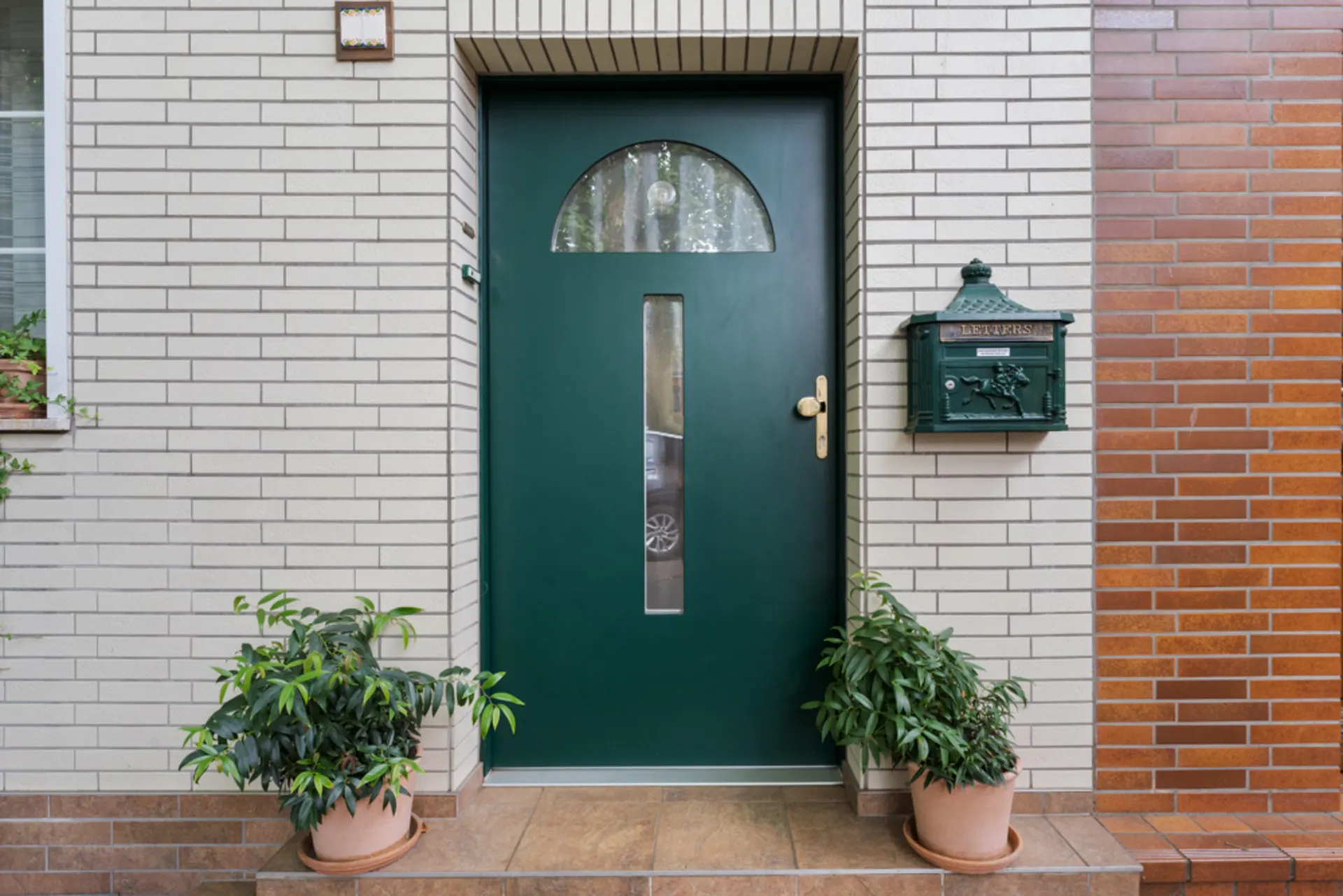 Grüne Haustür mit Briefkasten und zwei Pflanzen in Töpfen auf gefliestem Eingang.