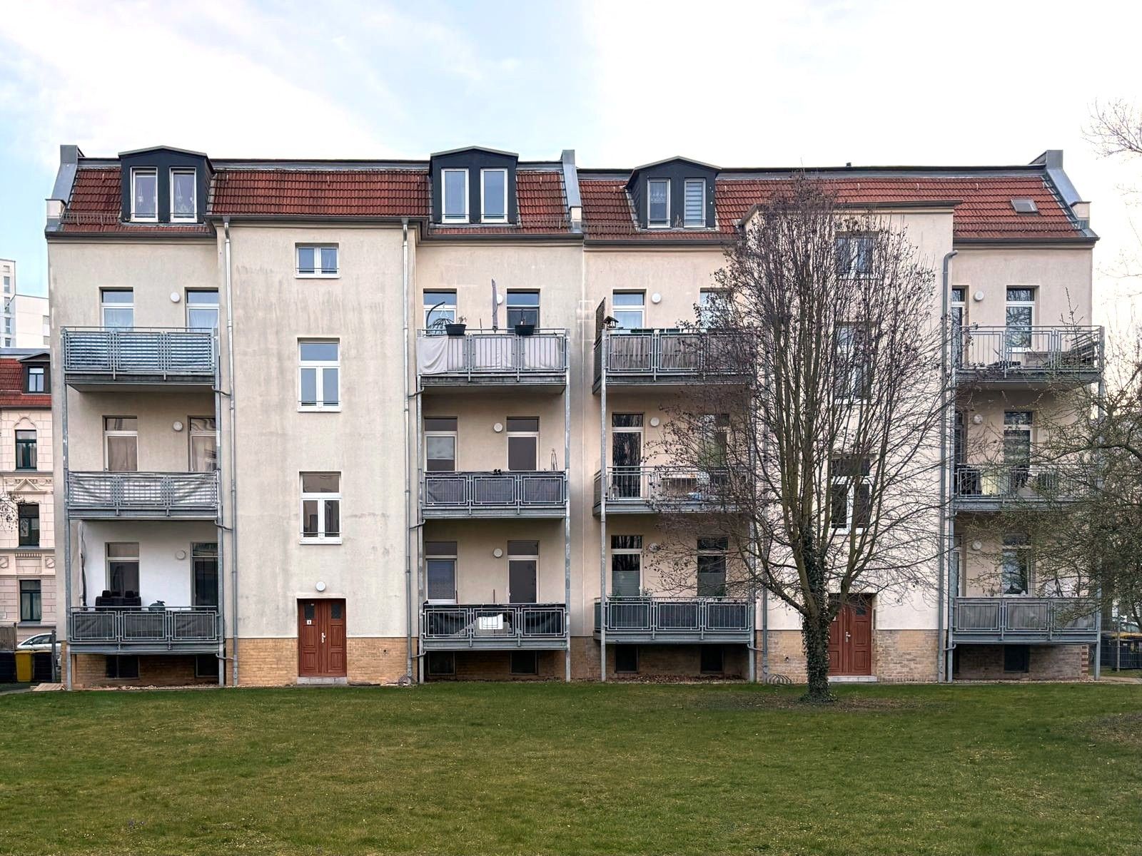 Mehrstöckiges Wohnhaus mit Balkonen und roten Dachfenstern, davor eine Wiese mit Baum.