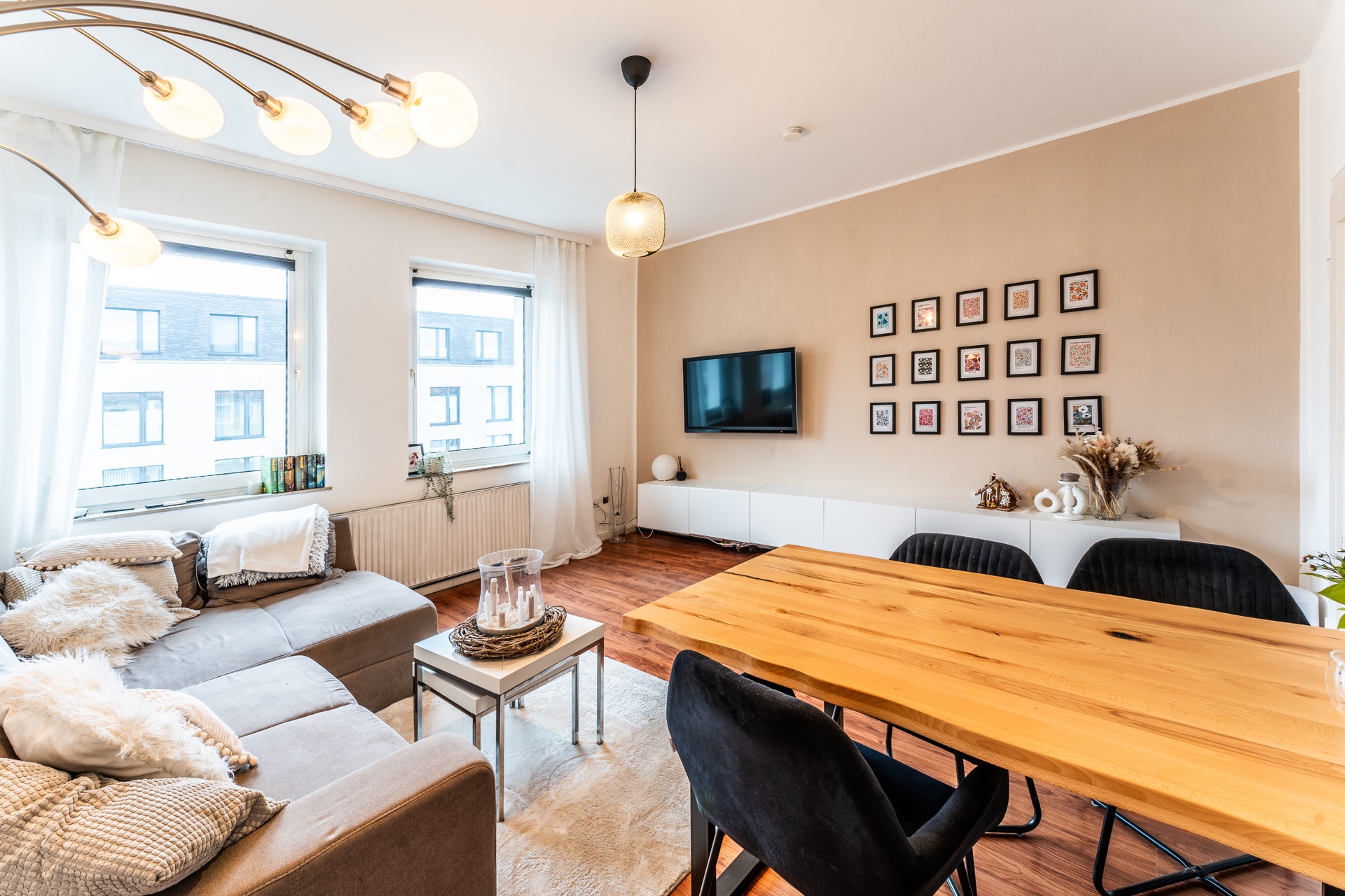 Modernes Wohnzimmer mit Holz-Esstisch, grauem Sofa, Wand-TV und großer Fensterfront.