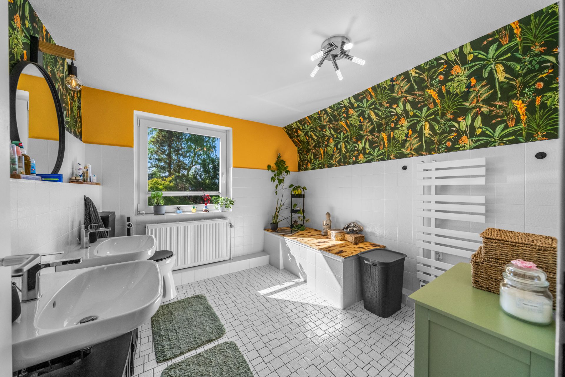 Helles Badezimmer mit Badewanne, zwei Waschbecken, großem Fenster und bunter Pflanzen-Tapete.
