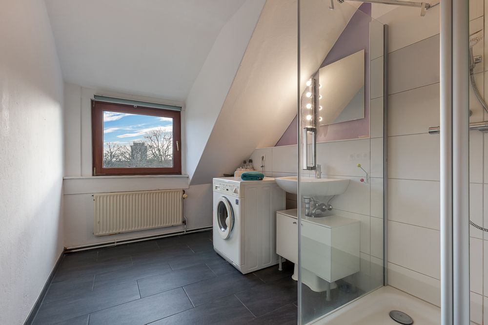 Modernes Bad mit Waschmaschine, Glasdusche, Waschbecken und Fenster unter einer Schräge.