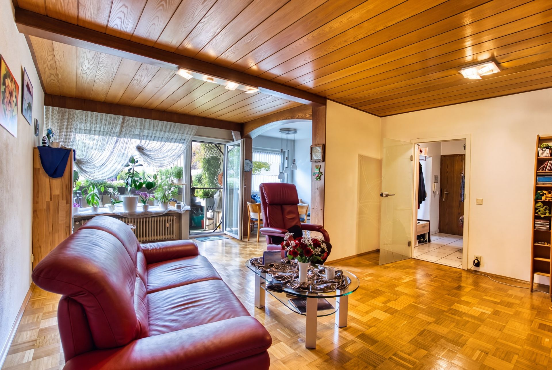 Wohnzimmer mit rotem Ledersofa, Glastisch, Holzdecke und Zugang zum Balkon.