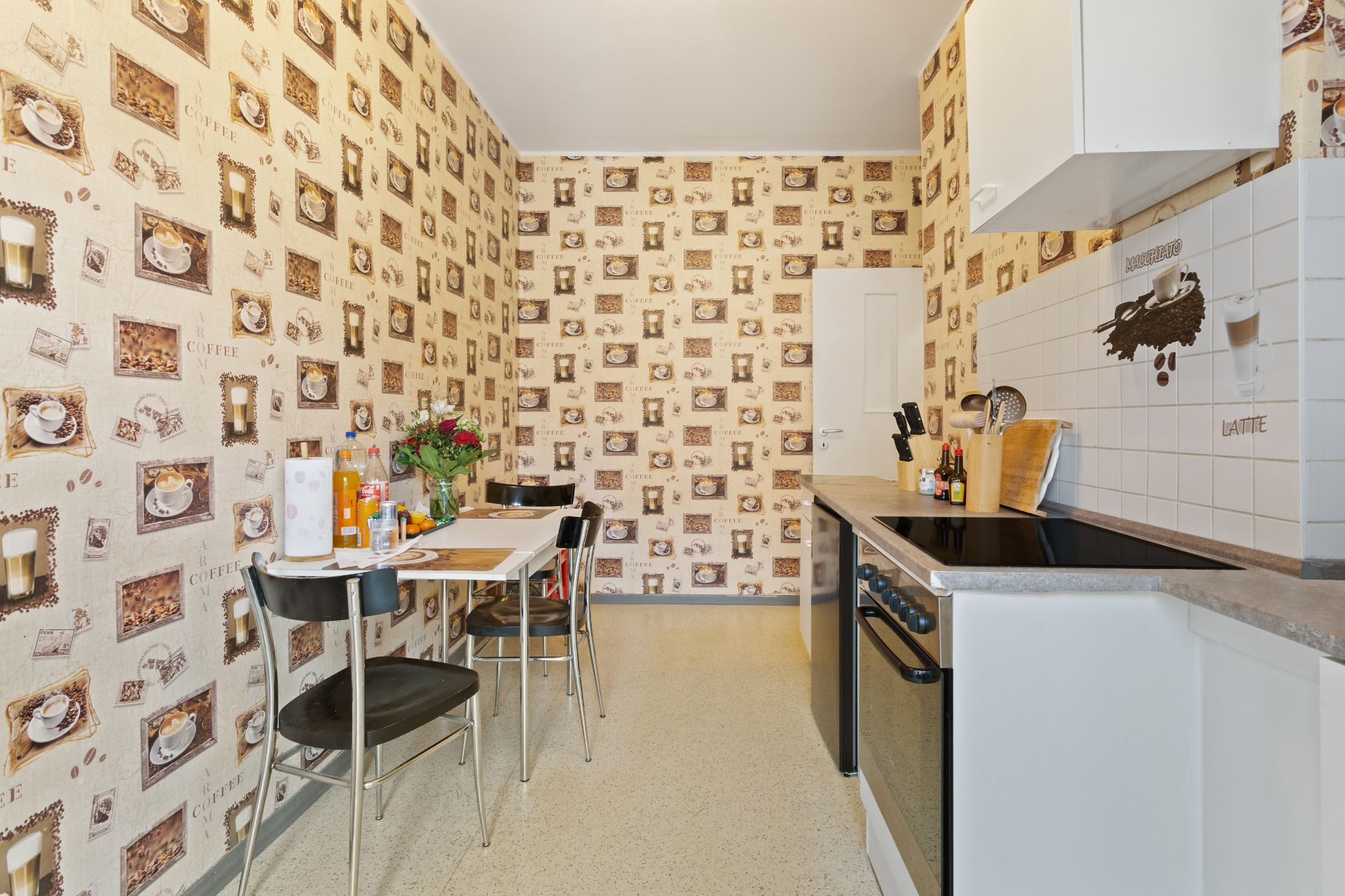 Schmale Küche mit Kaffeemotiven an der Wand, Esstisch mit Stühlen und Elektroherd.