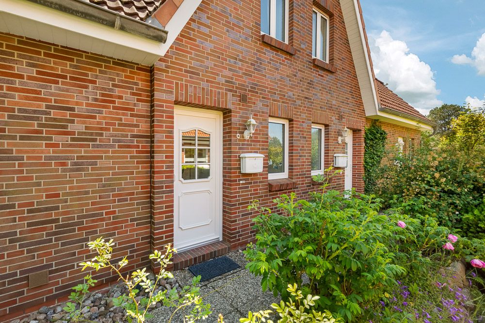 Backsteinfassade eines Hauses mit weißer Tür, Fenstern und Vorgarten mit Blüten.