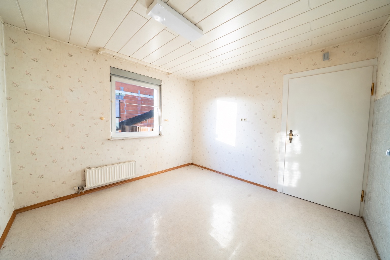 Helles, leeres Zimmer mit Fenster, Tür, Heizkörper und gemusterten Tapeten.