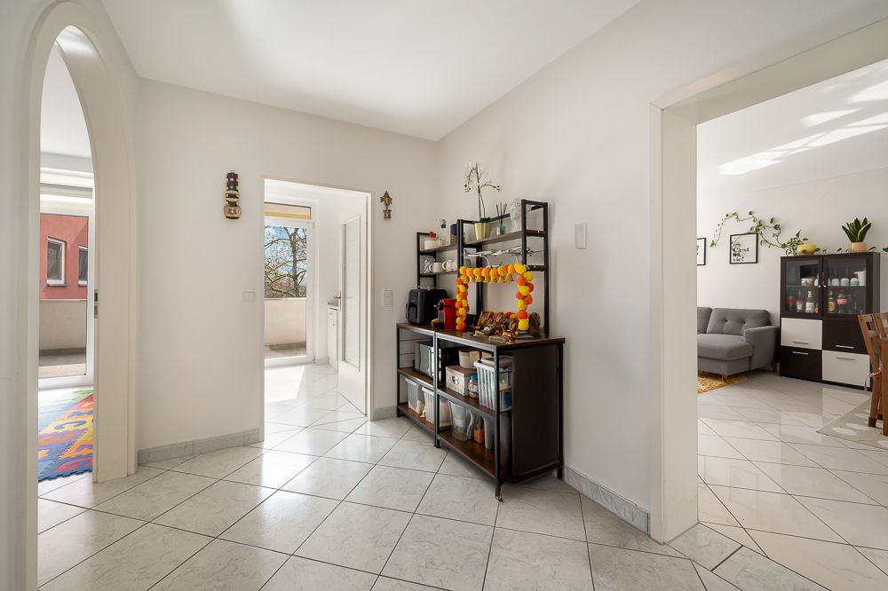 Heller Flur mit Fliesenboden, Regal und Blick in ein Wohnzimmer mit Sofa und Schrank.