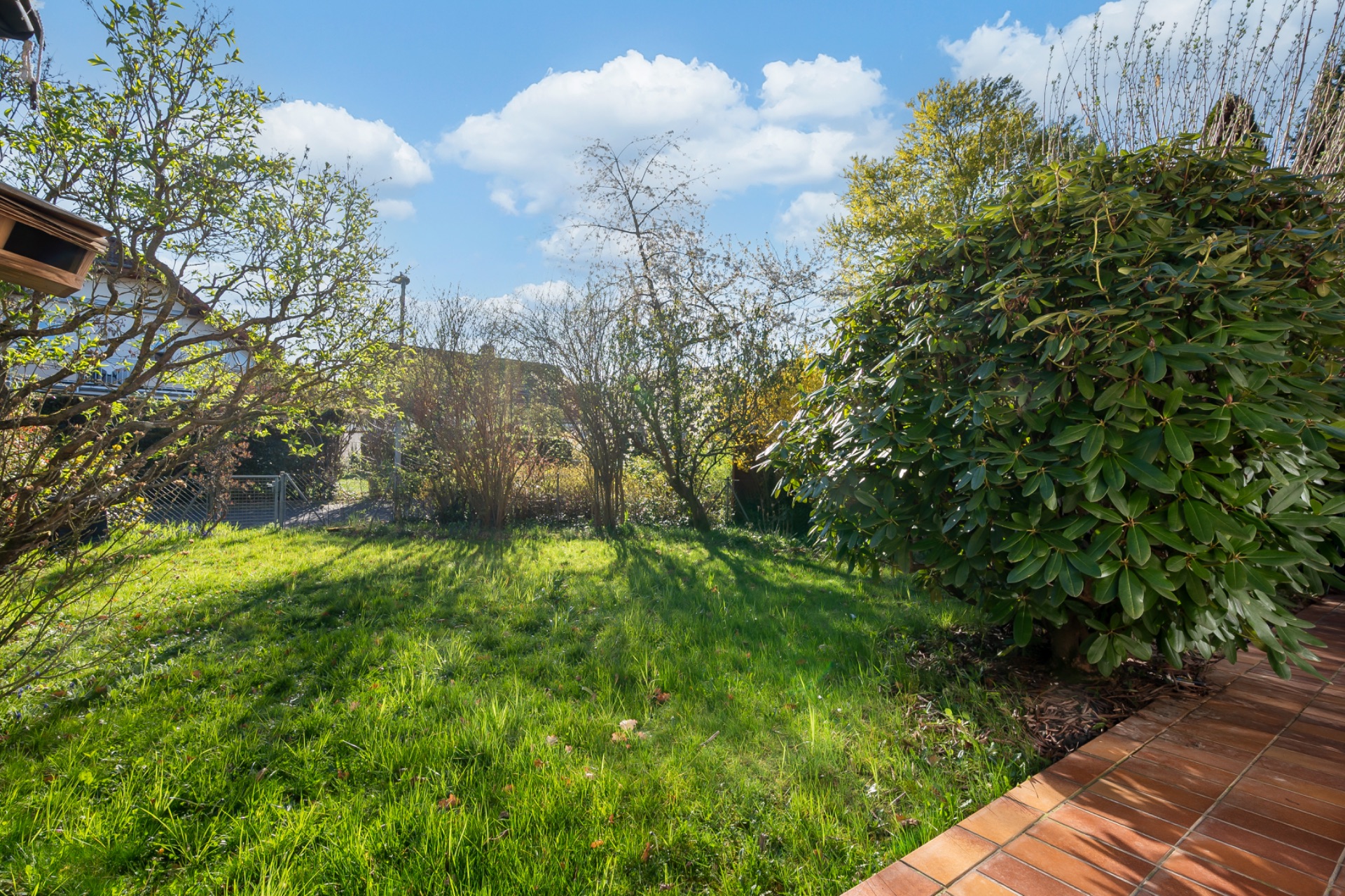 Garten mit grünem Rasen, Büschen und Bäumen, daneben ein Terrassenbereich mit roten Fliesen.