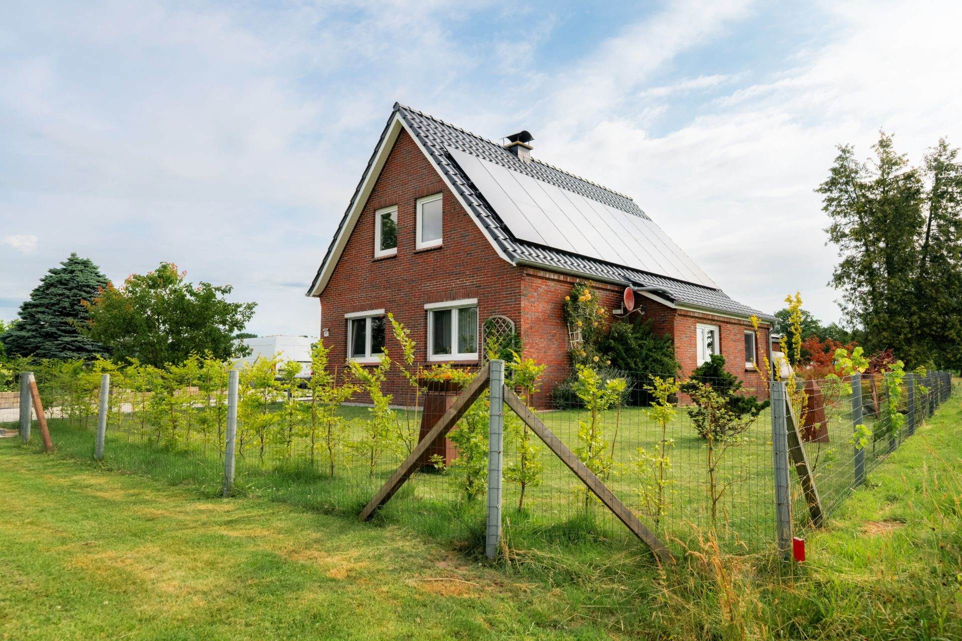 Backsteinwohnhaus mit Solarmodulen auf dem Dach, umgeben von Garten und Zaun.