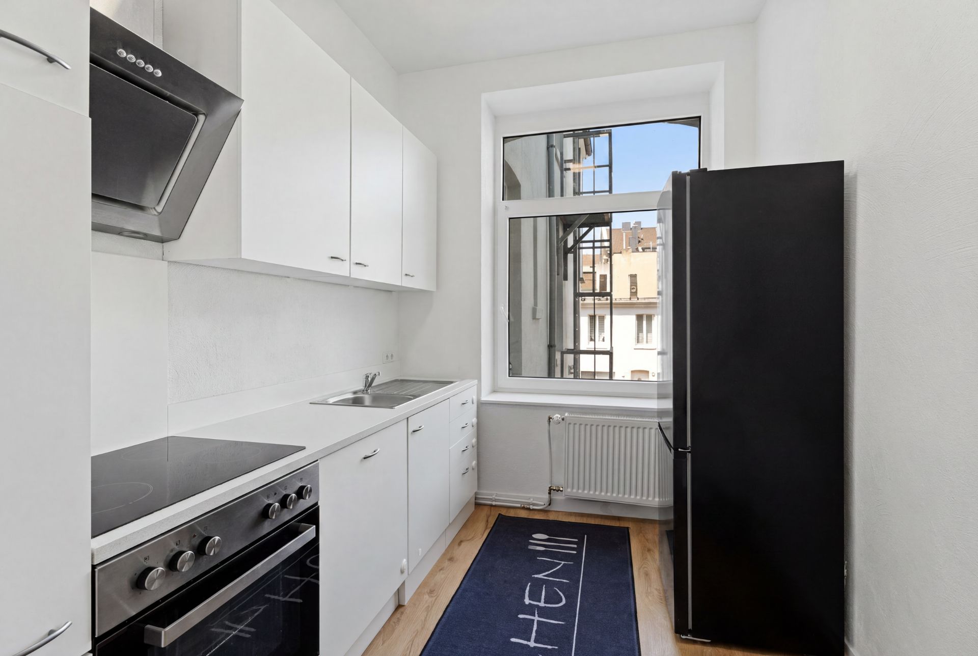 Schmale, moderne Küche mit weißen Schränken, Herd, Spüle, schwarzem Kühlschrank und Fenster.