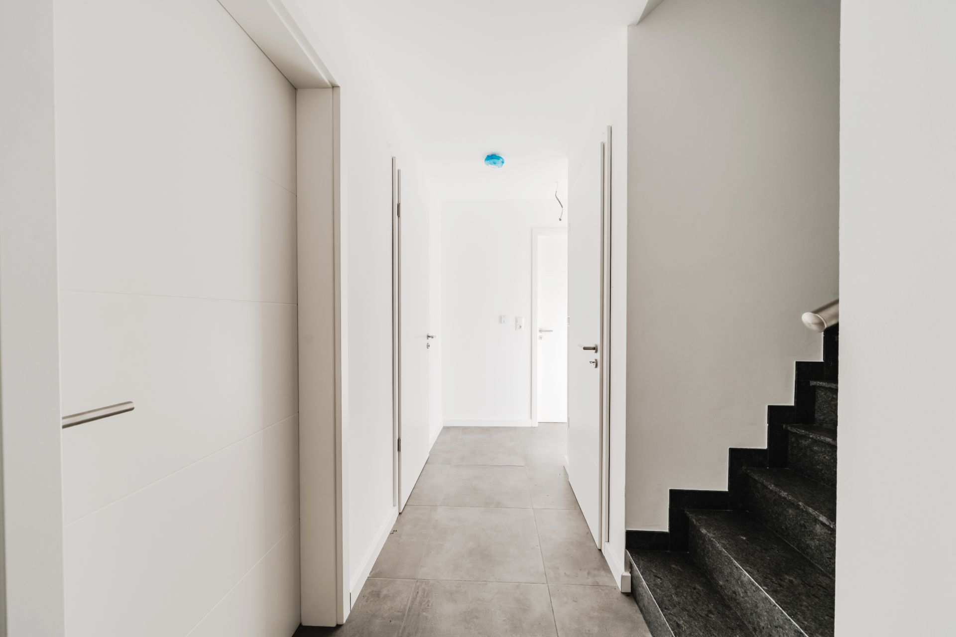 Heller Flur mit grauem Boden, mehreren Türen und einer schwarzen Treppe rechts.