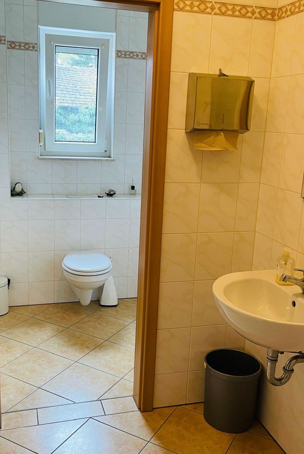 Gefliestes Badezimmer mit Waschbecken, Mülleimer, Papierhandtuchspender und Toilette am Fenster.