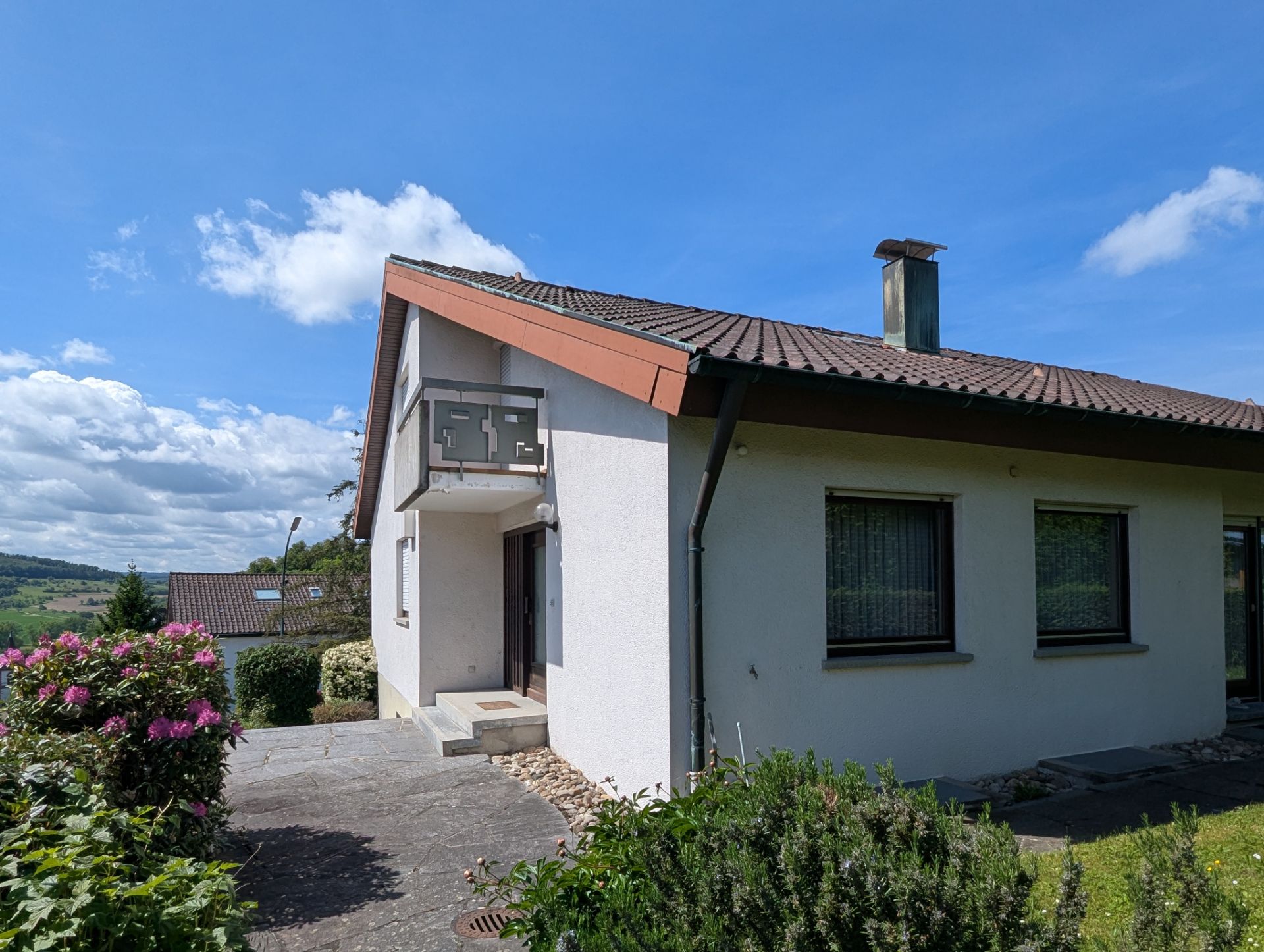 Einfamilienhaus mit hellem Putz, kleinem Balkon und Garten bei sonnigem Wetter.