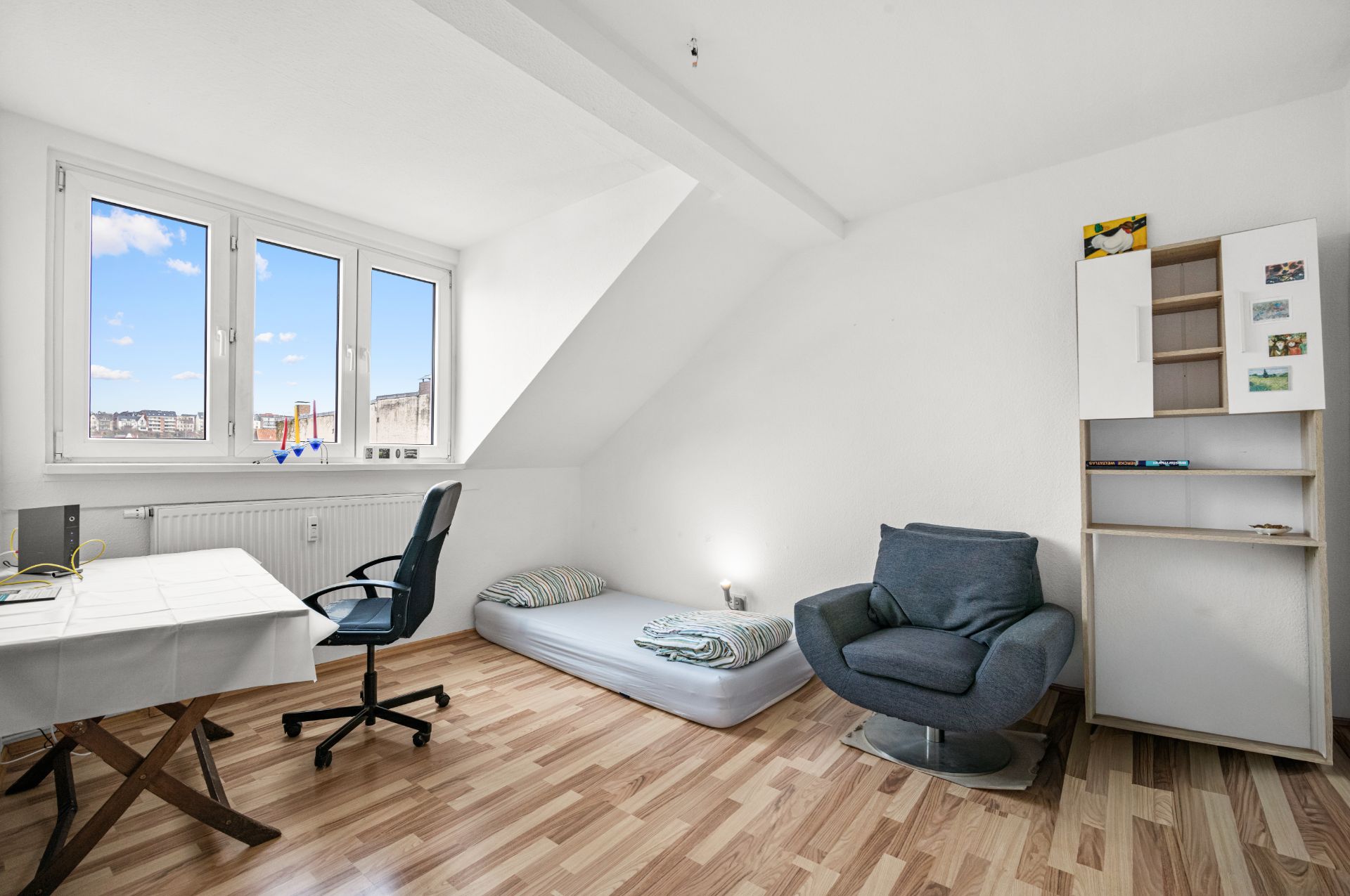 Helles Zimmer mit Schreibtisch, Stuhl, Matratze auf dem Boden, Sessel und Regal auf Laminatboden.
