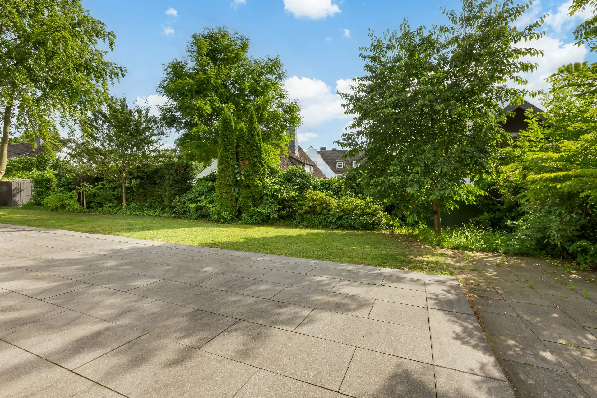 Gepflasterte Terrasse mit Blick auf einen grünen Garten und Bäume an einem sonnigen Tag.