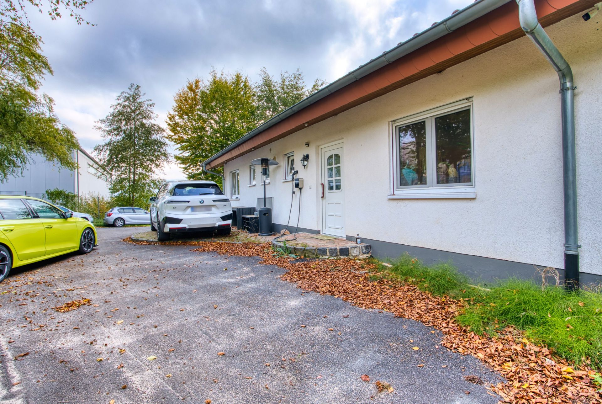 Haus mit weißer Fassade, Haustür, Fenster und drei geparkten Autos auf Einfahrt im Herbst.