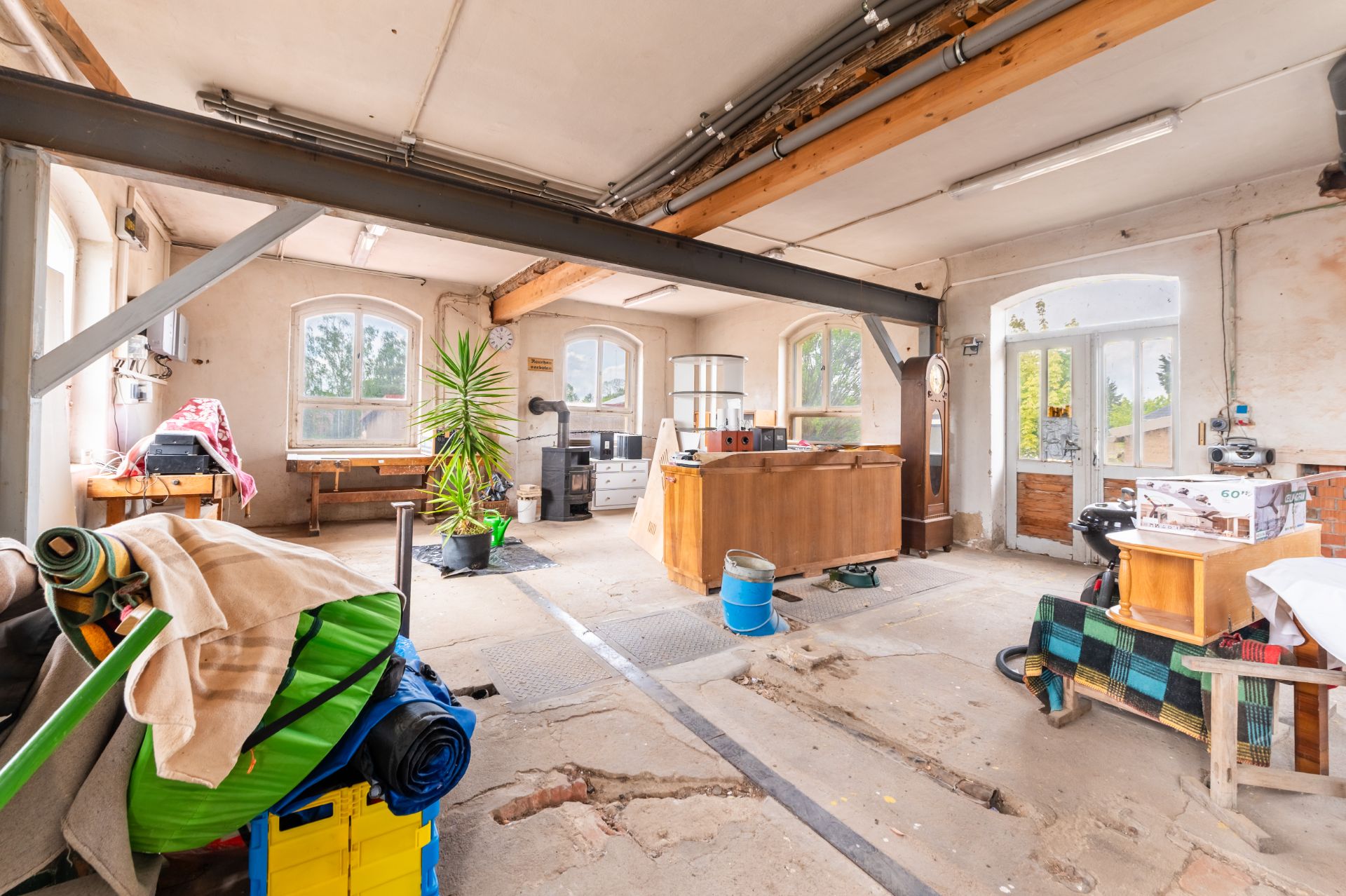 Heller Raum mit sichtbaren Holzbalken, Pflanzen, Tischen und verschiedenen Gegenständen auf dem Boden.