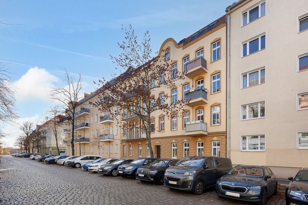 Mehrfamilienhaus mit Balkonen an einer Pflasterstraße, parkende Autos und kahle Bäume im Vordergrund.