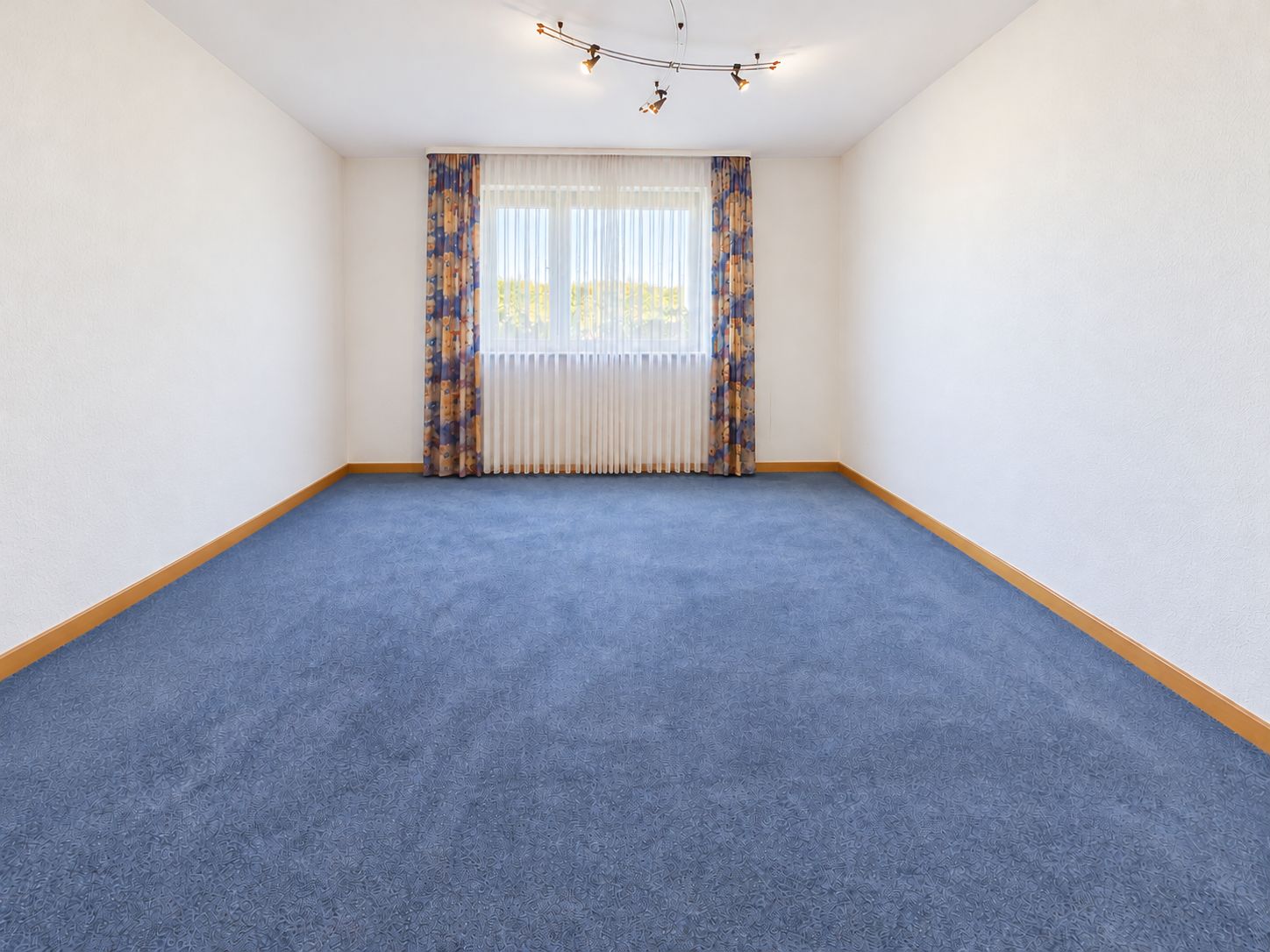 Leeres Zimmer mit blauem Teppich, weißen Wänden und Fenster mit Vorhängen.