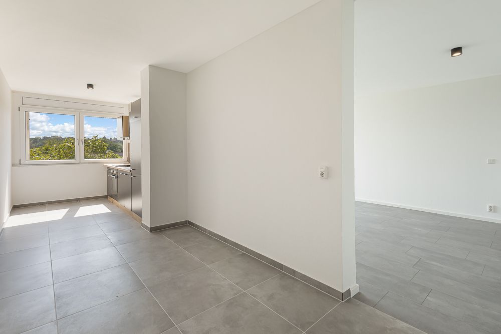 Helle, leere Küche mit großem Fenster und grauem Fliesenboden, angrenzend an einen leeren Wohnraum.