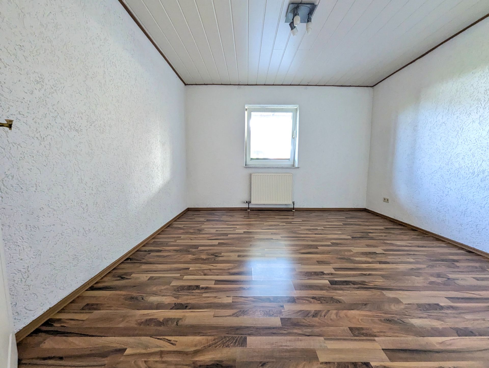 Leerer Raum mit weißer Decke, Holzfußboden und einem Fenster mit Heizung darunter.