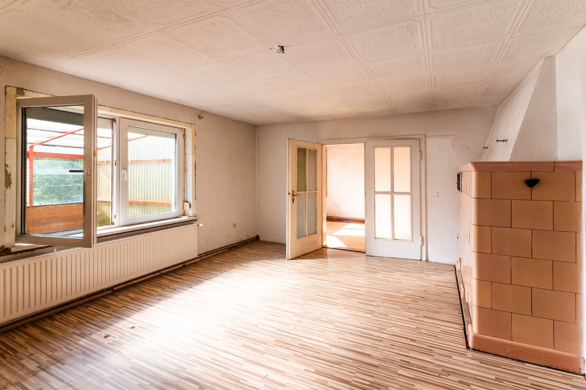 Heller leerer Raum mit offenem Fenster, Holzboden und braunem Kachelofen.