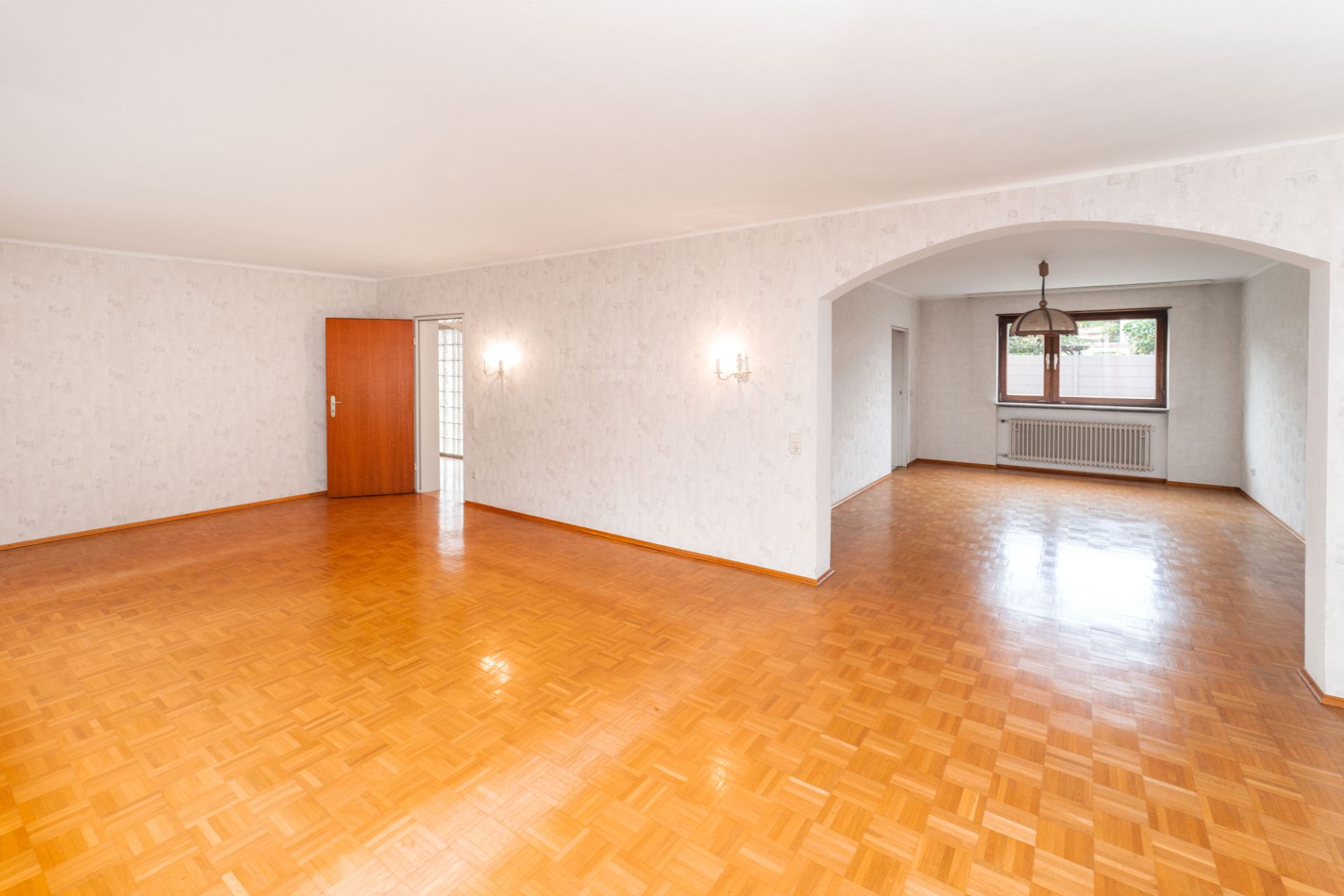 Helles Wohnzimmer mit Parkettboden, weißen Wänden und großem Fenster, verbunden mit Esszimmer durch Rundbogen.