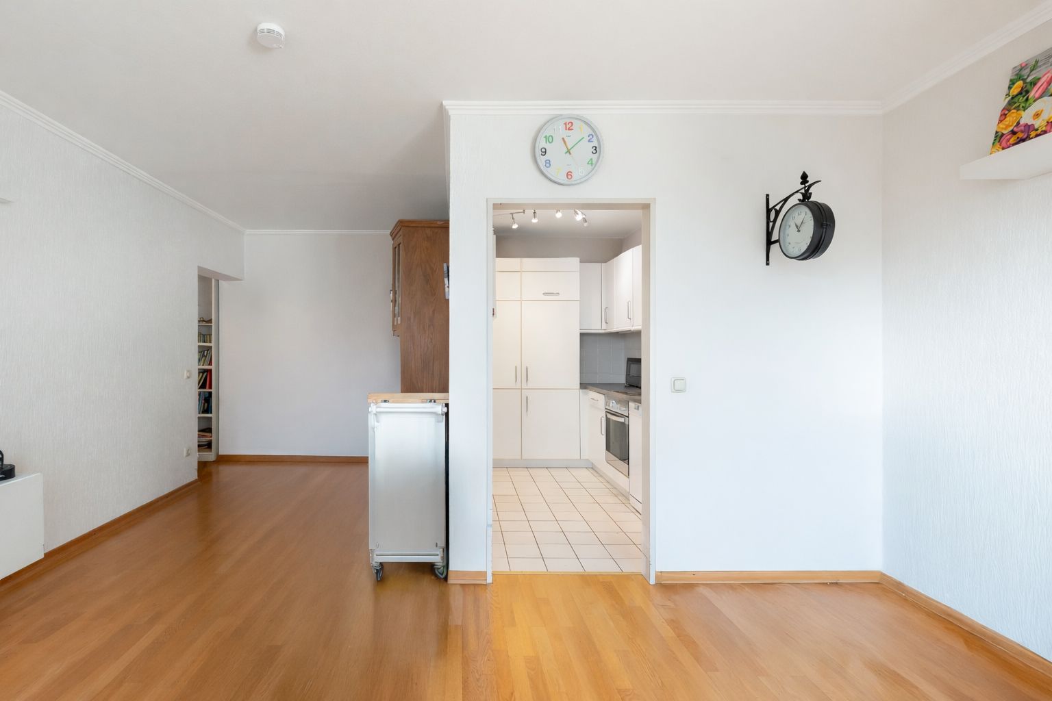 Helles Wohnzimmer mit Holzboden und Blick in eine weiße Küche mit Fliesenboden.