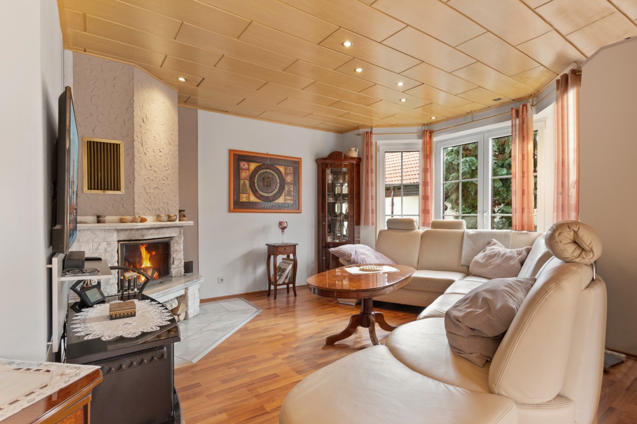 Wohnzimmer mit Kamin, beiger Ledercouch, Holztisch und großen Fenstern mit Vorhängen.