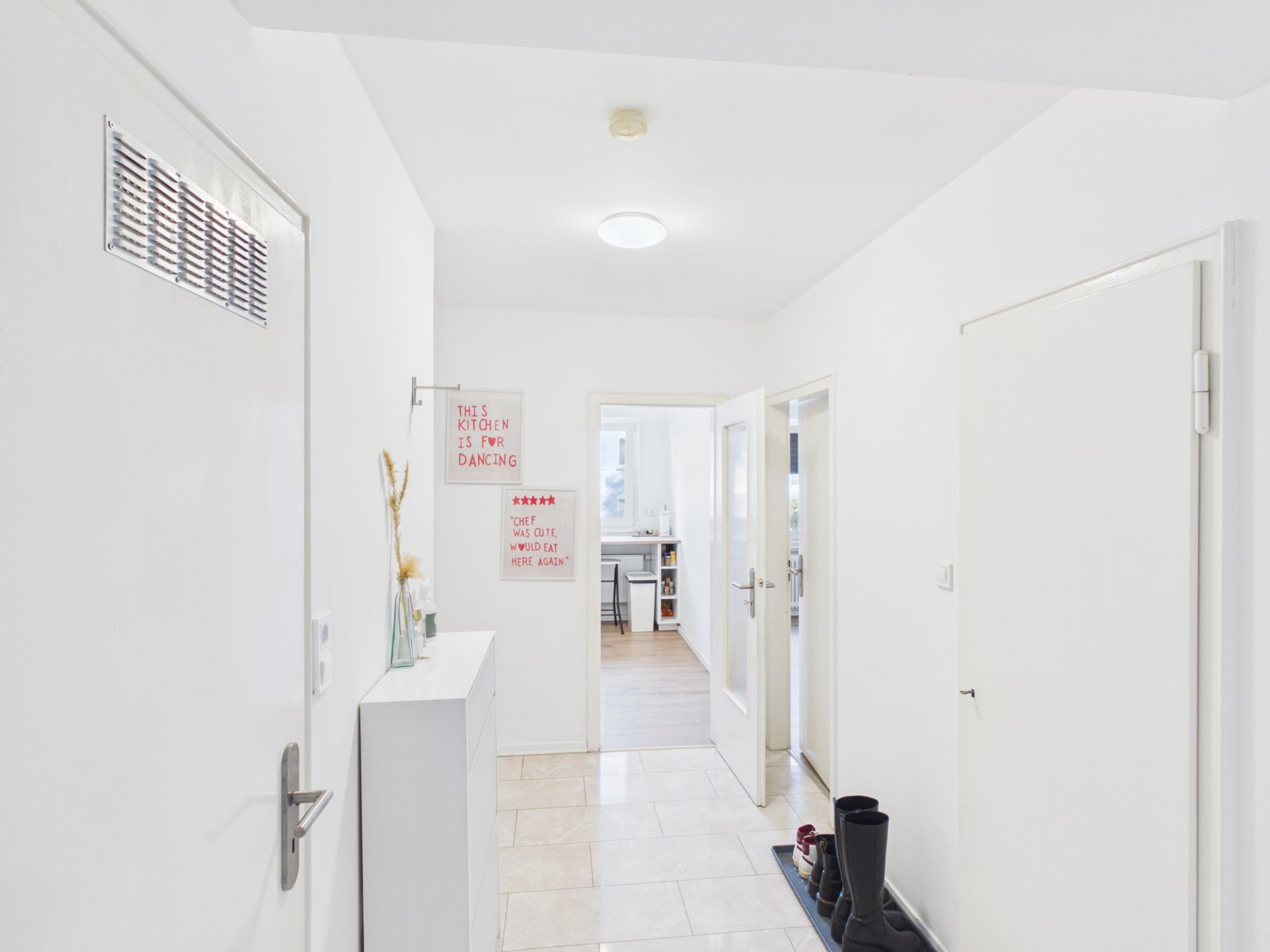 Heller Flur mit weißer Wand, Schuhablage und geöffneter Tür zum nächsten Raum.