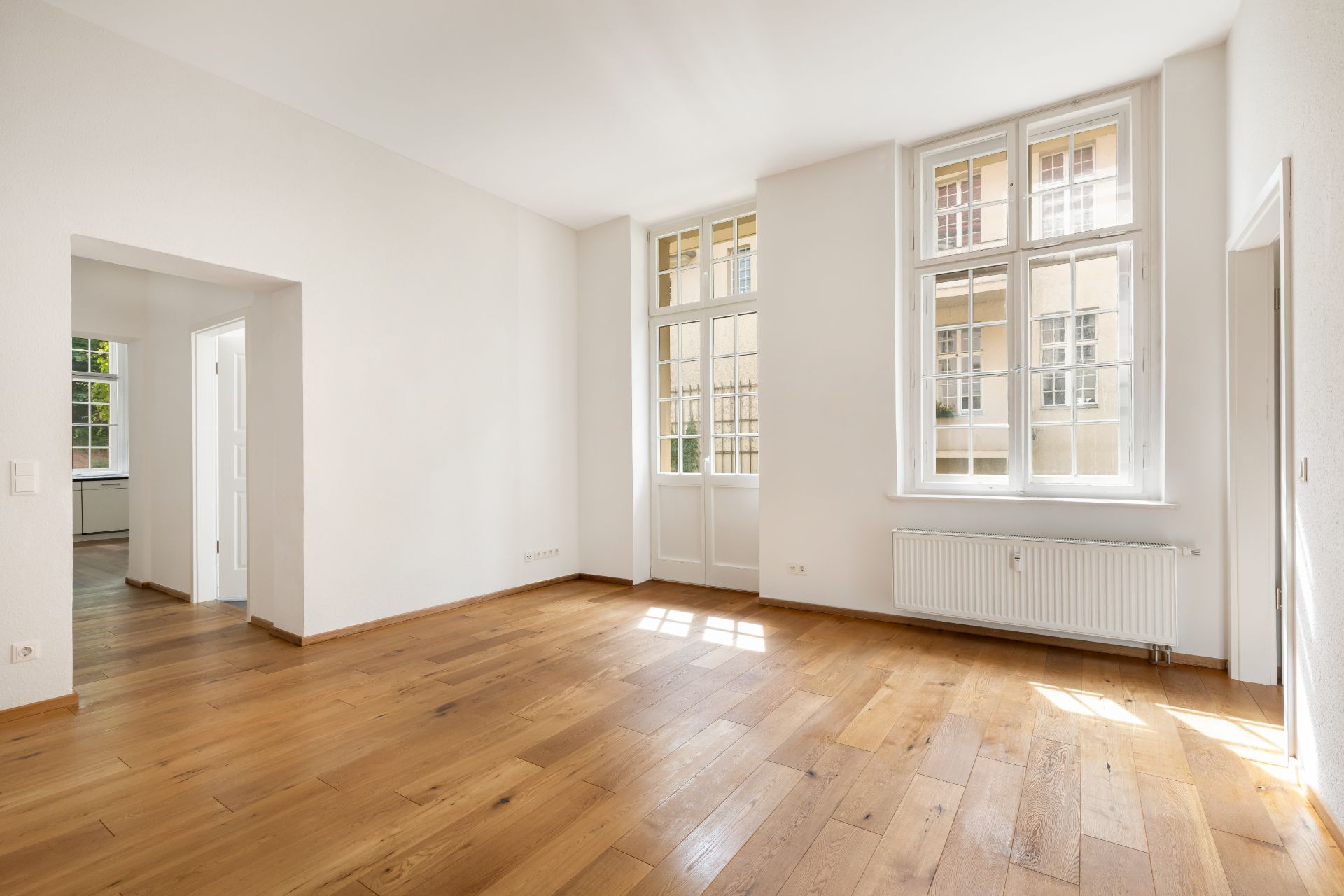 Helles leeres Zimmer mit Holzboden, weißen Wänden und großen Fenstern.