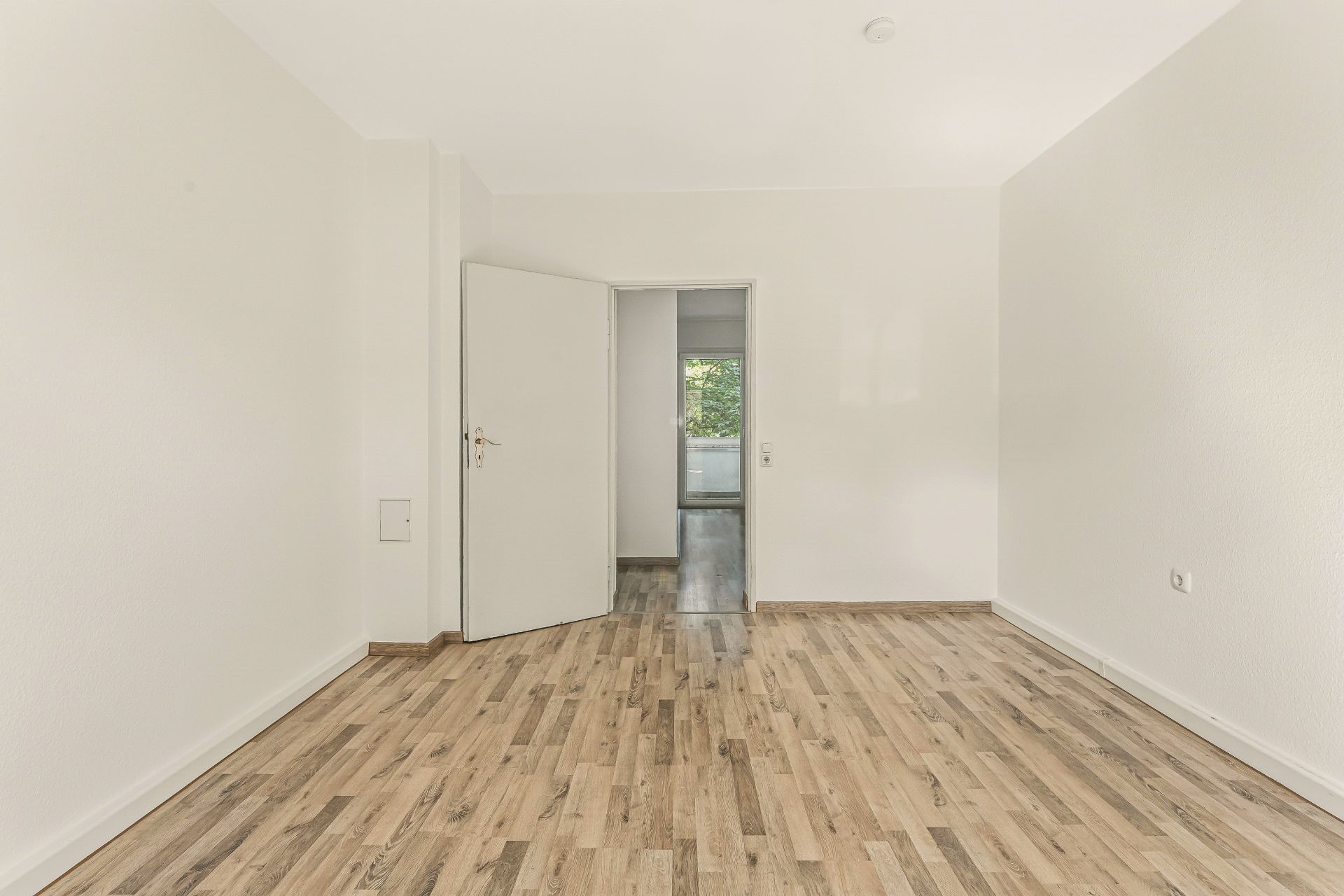 Leerer Raum mit weiß gestrichenen Wänden, Holzboden und offener Tür zu einem weiteren Zimmer.